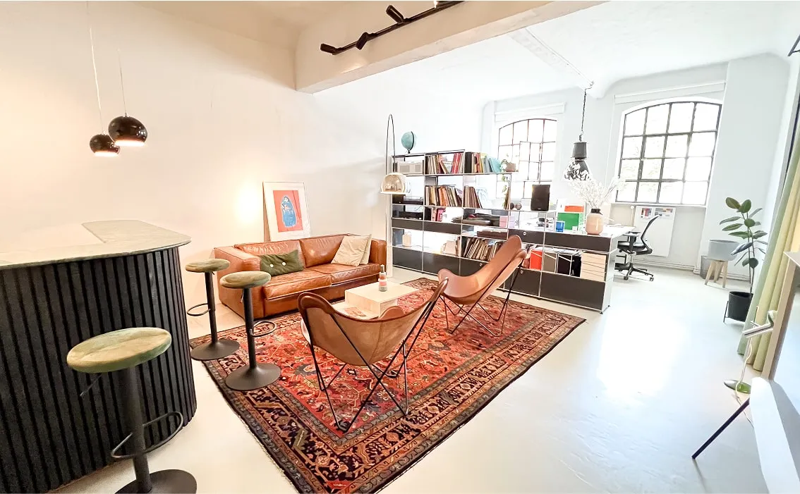 Lounge- und Arbeitsbereich – Großer heller Raum mit Teppich, Sesseln, Schreibtisch und Regalwand.