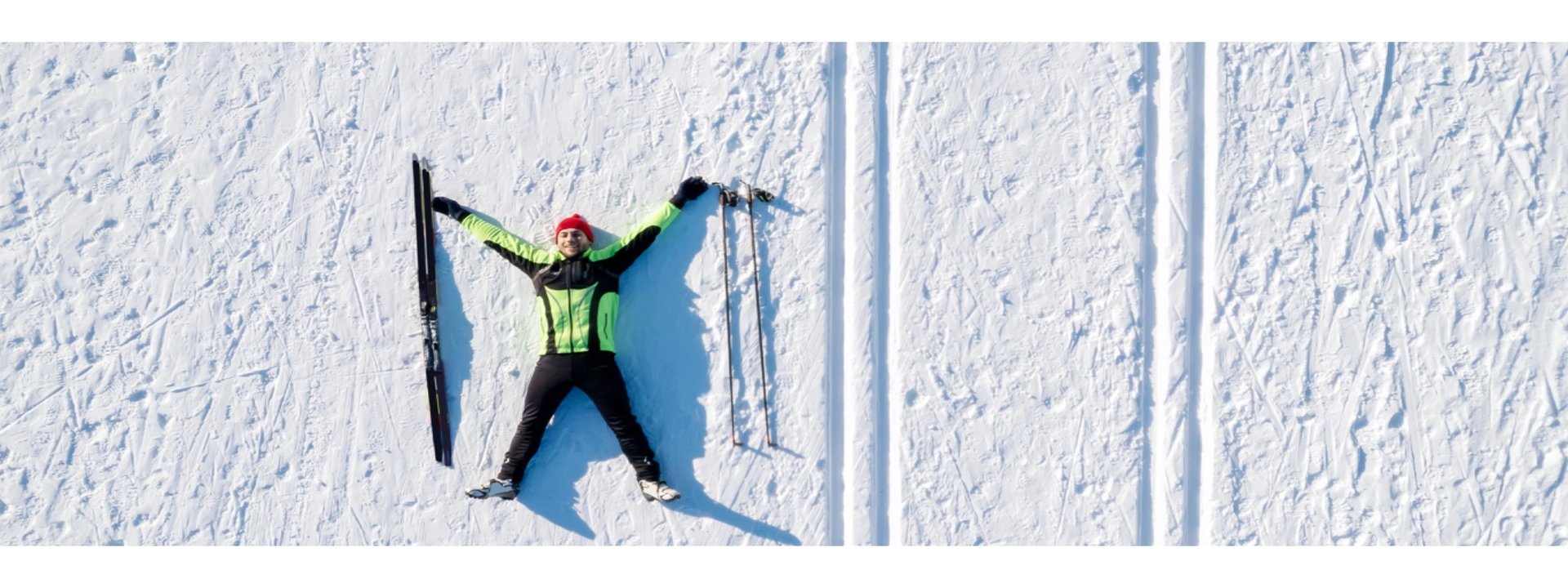 Smiling cross-country skier lying on snow with skis and poles beside him, aerial view of winter landscape in Finnish Lapland.