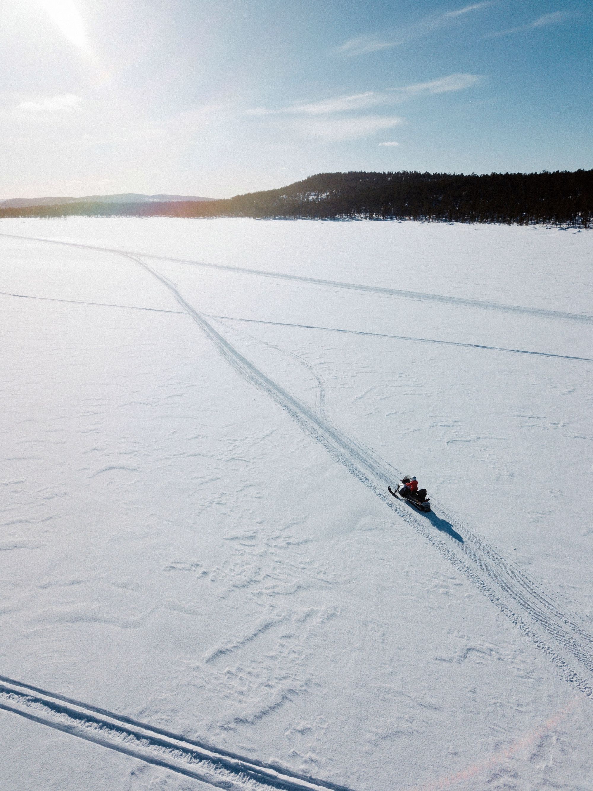 Aerial view of a lone snowmobile crossing an open snowy landscape in Finnish Lapland, leaving peaceful tracks behind.