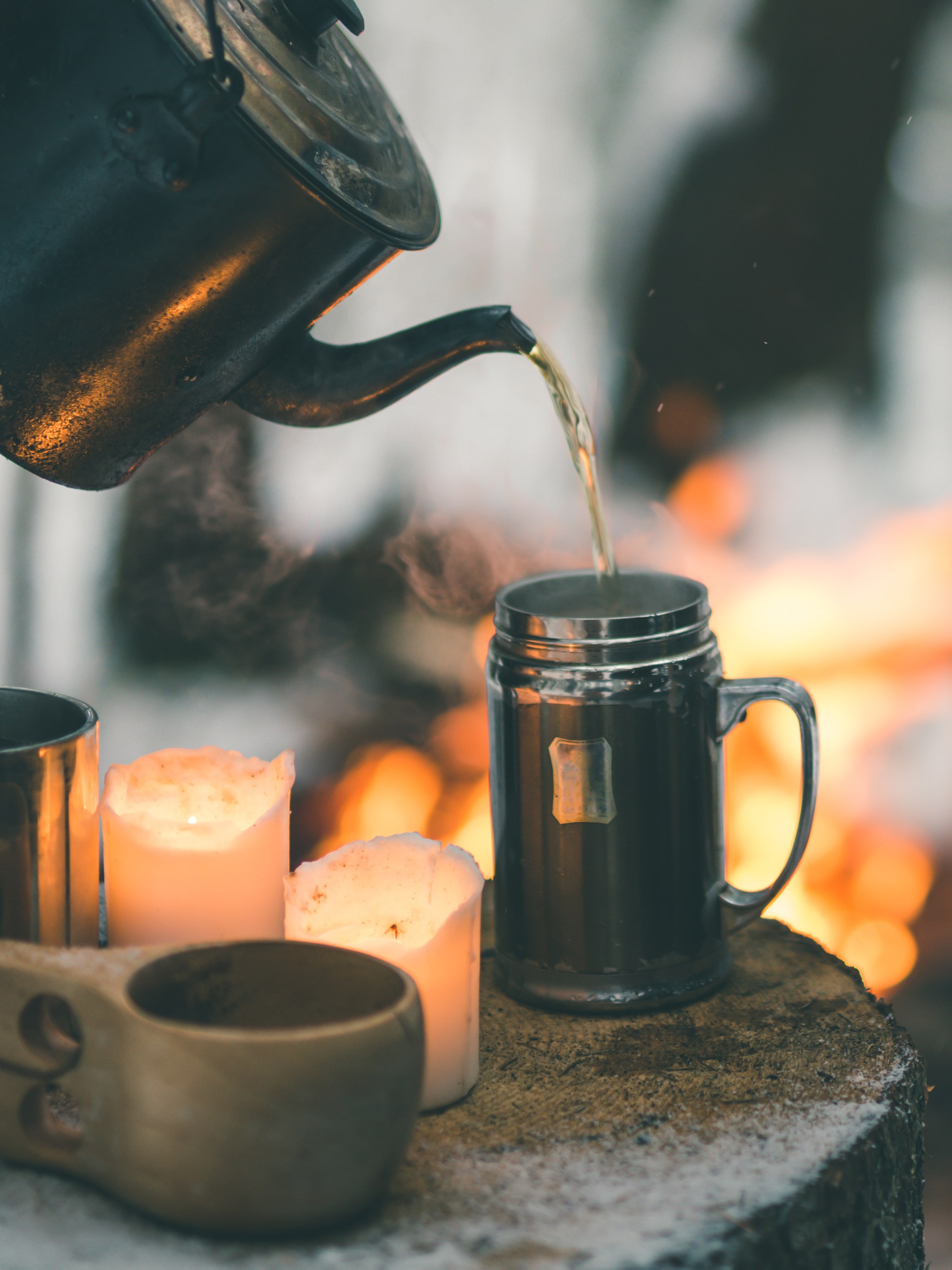 Tea being poured from a kettle into a campfire mug on a tree stump in a snowy Lapland forest.