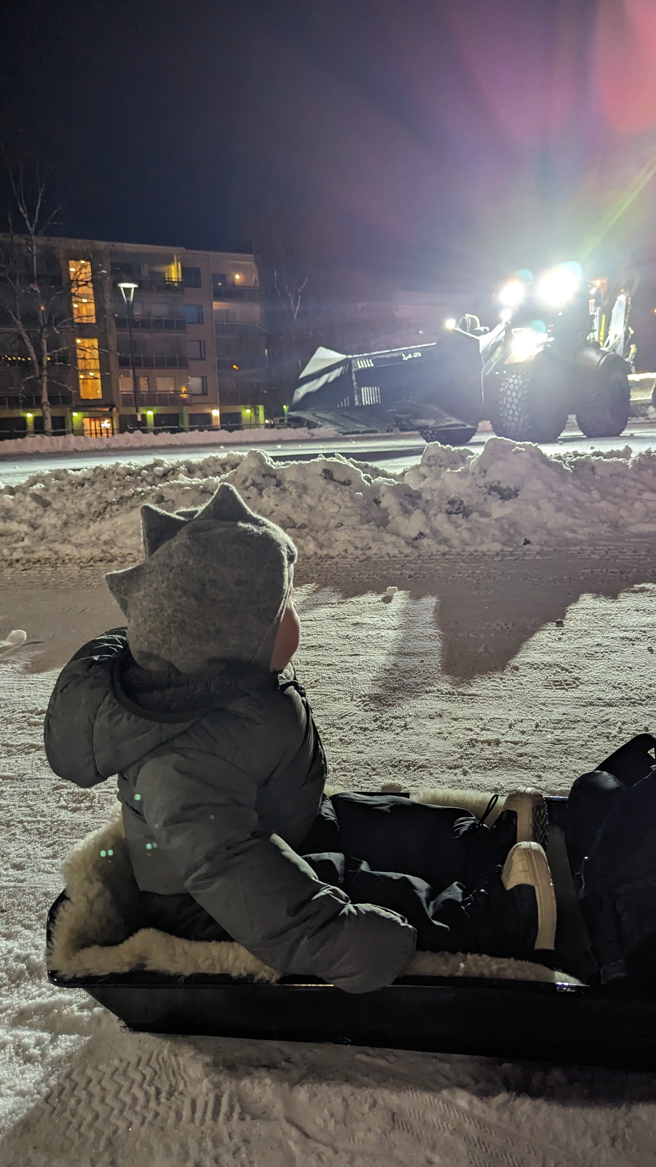 Little boy sitting in a sleigh on a dark winter evening in Rovaniemi, watching a snowplow clear the road.