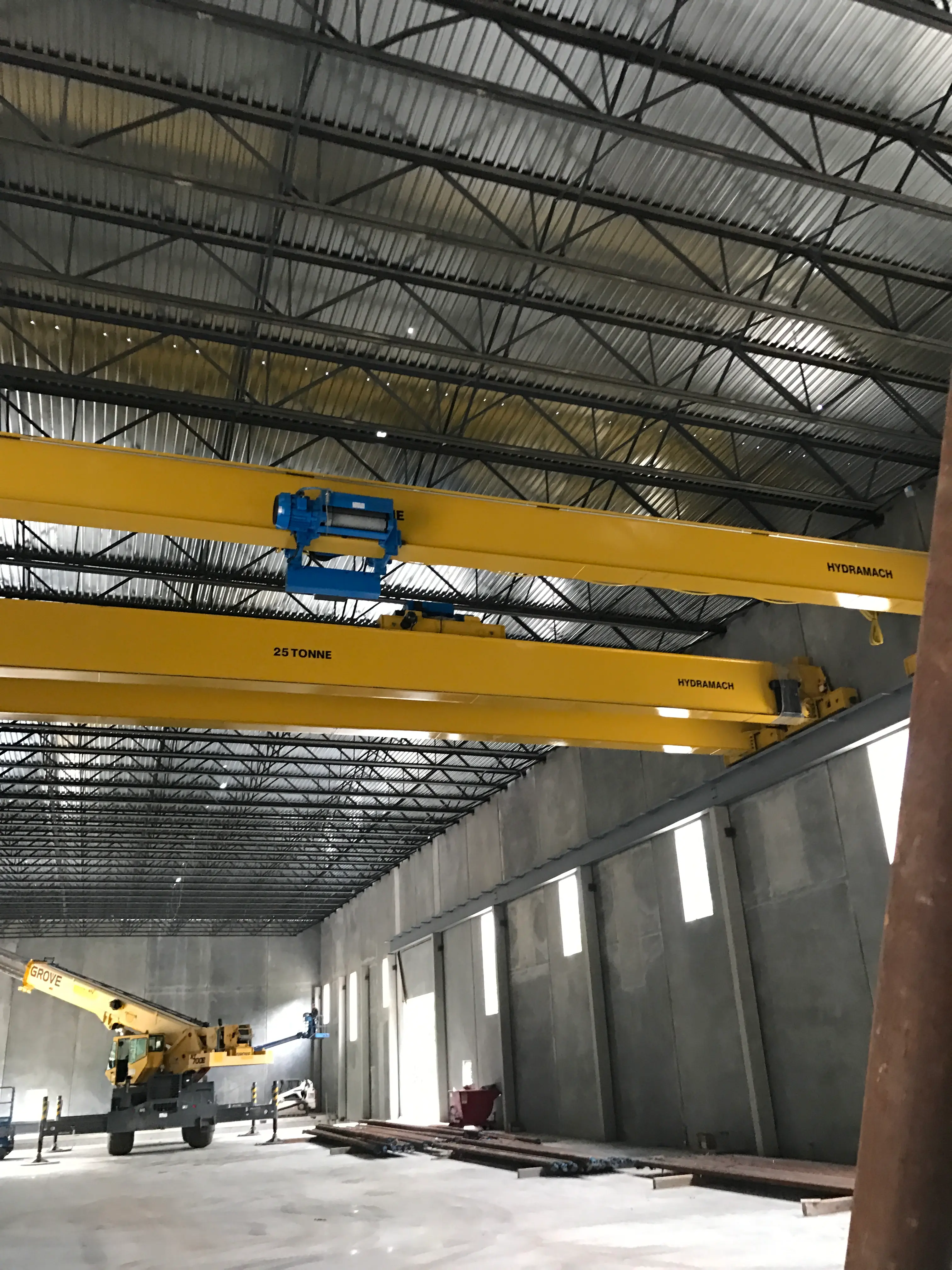 Interior view of an industrial warehouse under construction, showing two large yellow overhead cranes labeled "25 Tonne" suspended from the ceiling, with a mobile crane positioned on the concrete floor below.