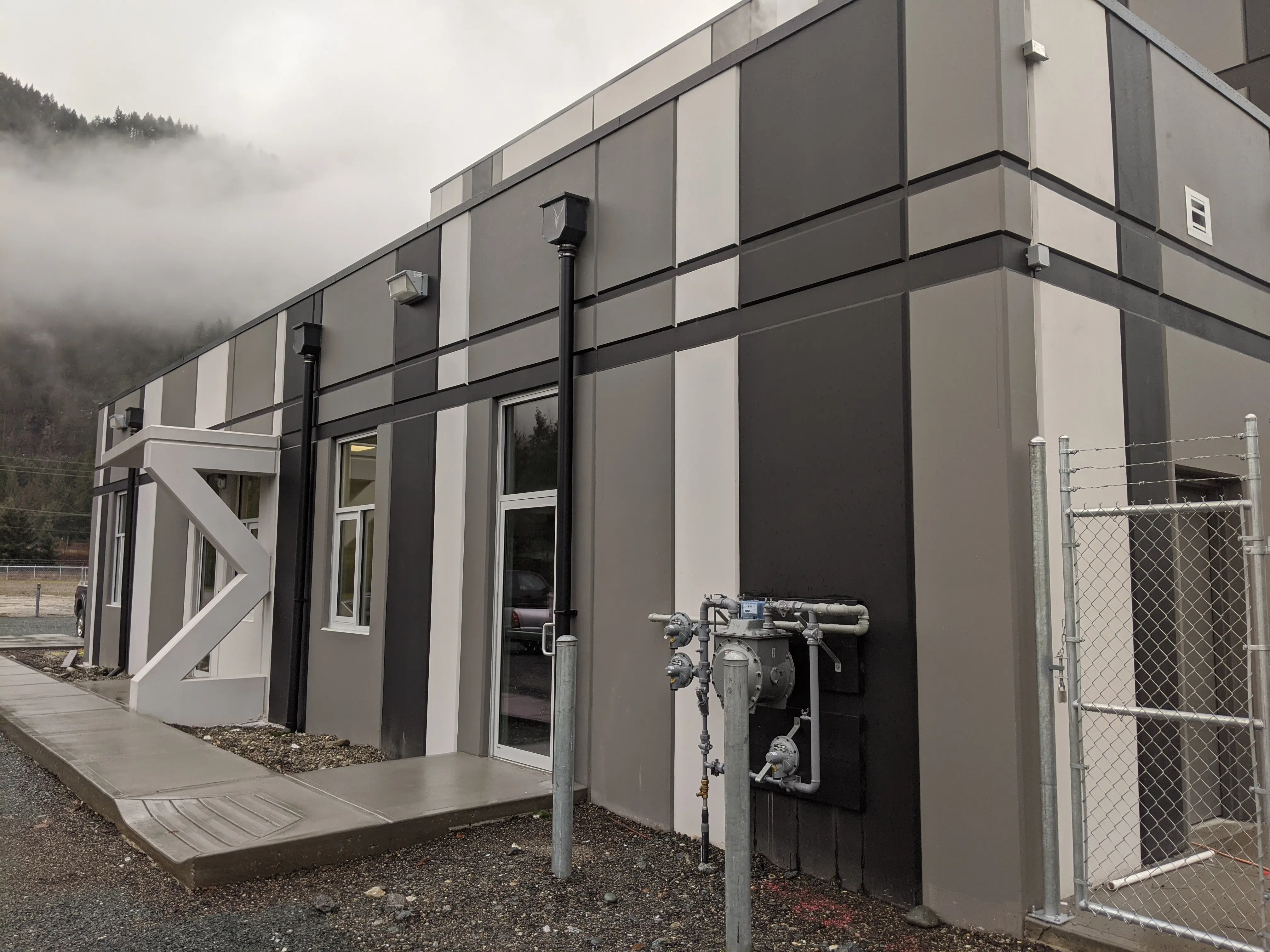 Exterior of a modern industrial building with a geometric design, featuring alternating gray and white vertical panels, multiple windows, and utility piping, set against a misty mountain backdrop.
