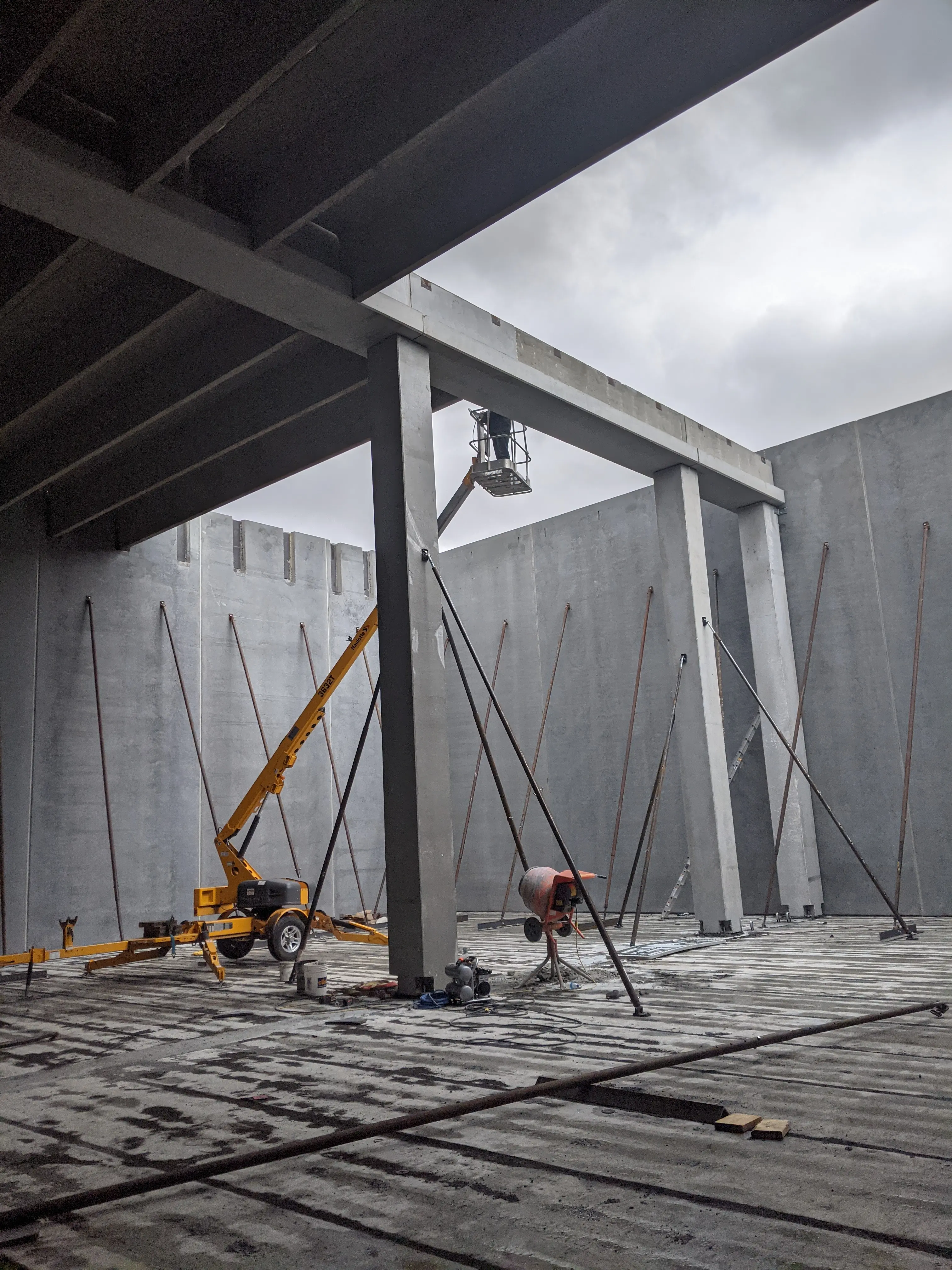 Large precast concrete structure under construction with support columns, steel braces, and workers on site using yellow construction equipment.