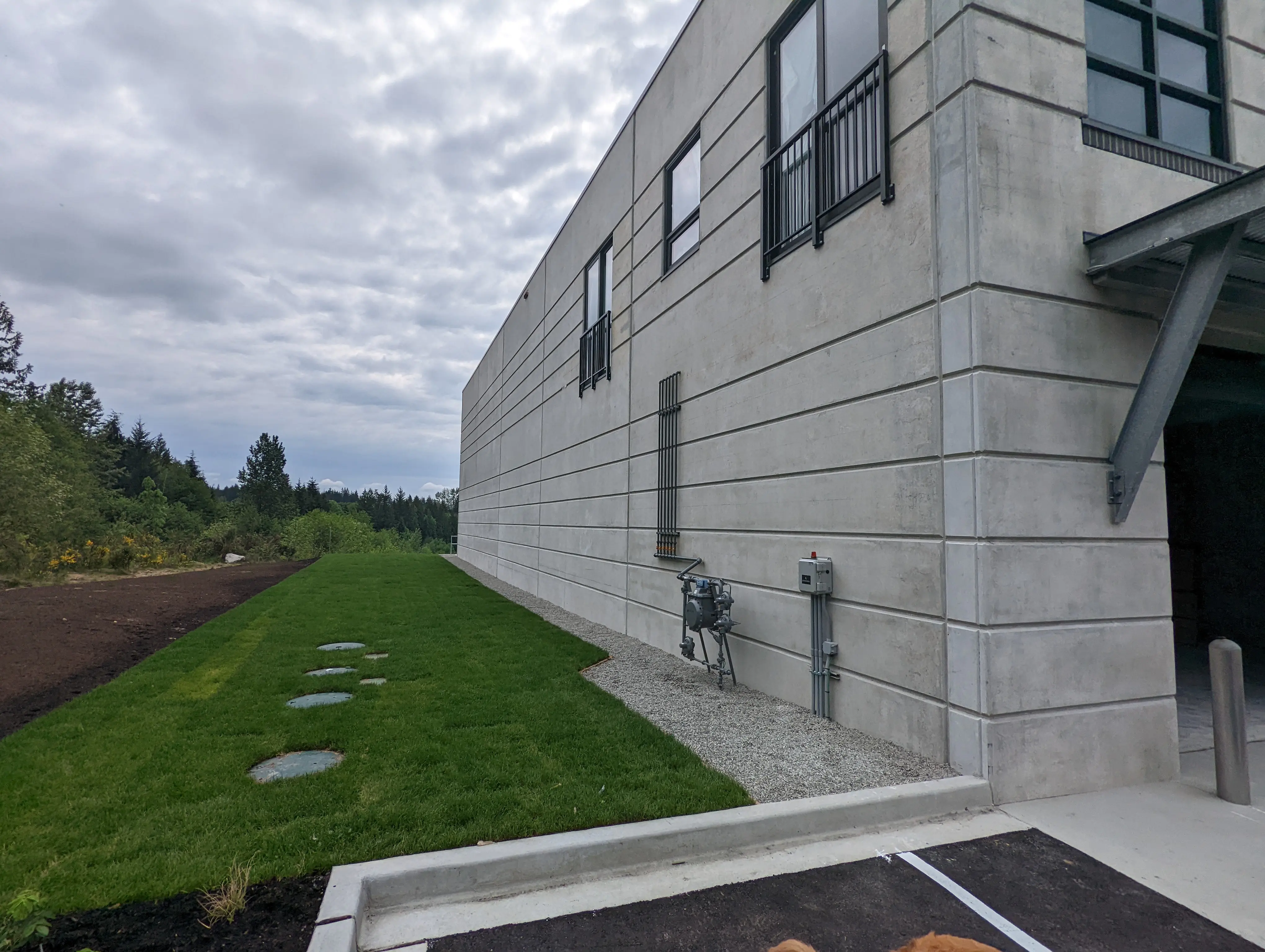 Exterior side view of a modern concrete industrial building with clean horizontal panel lines, surrounded by greenery and a neatly maintained lawn.