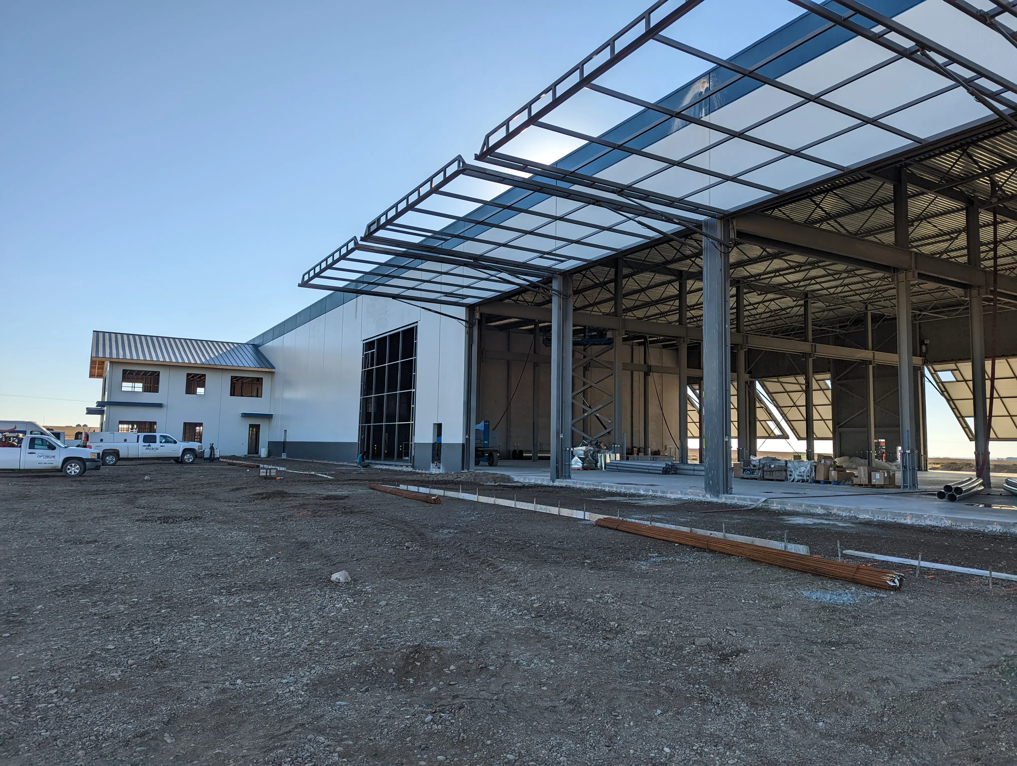 Exterior view of a large industrial building under construction with steel framework, partially installed roof panels, and utility trucks parked nearby.
