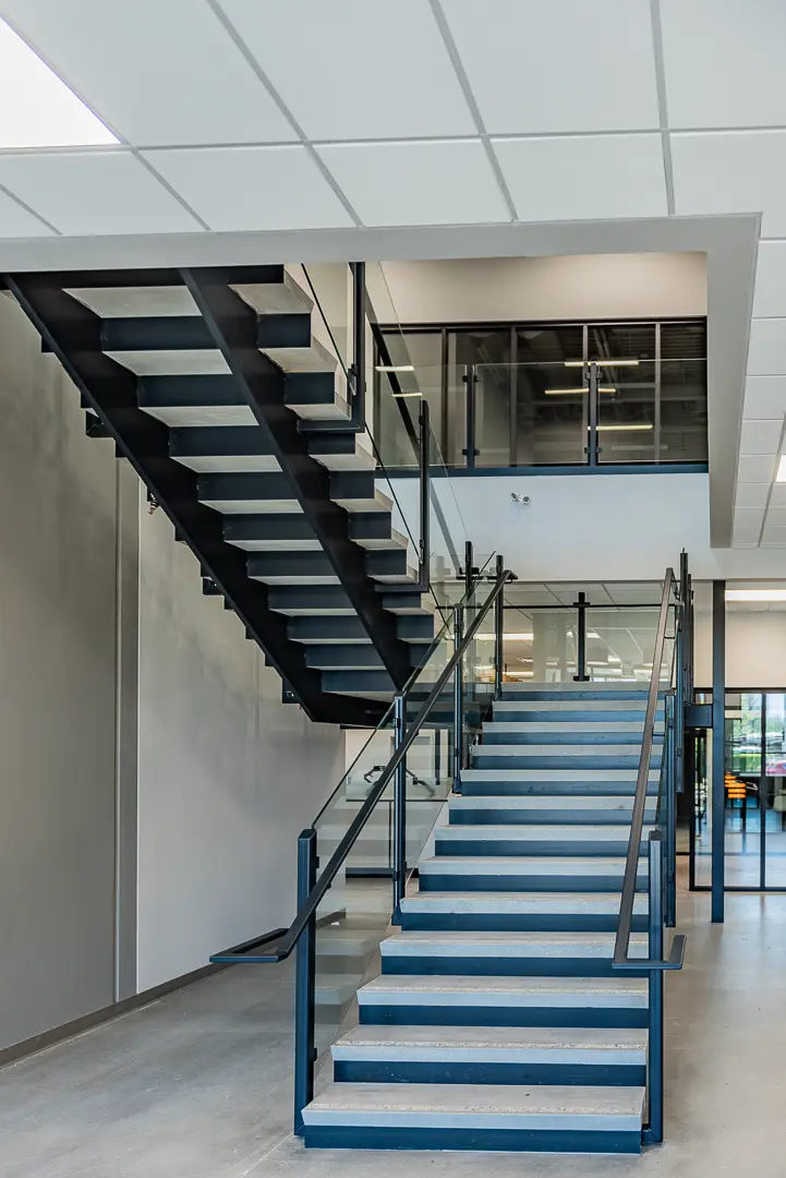 Modern interior staircase with black steel frame, blue steps, and glass railings leading to the second floor of an industrial office building.