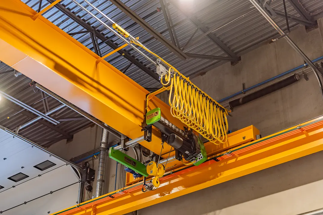 Close-up of a bright yellow overhead crane system with hoist and control cables suspended from the ceiling of a warehouse.