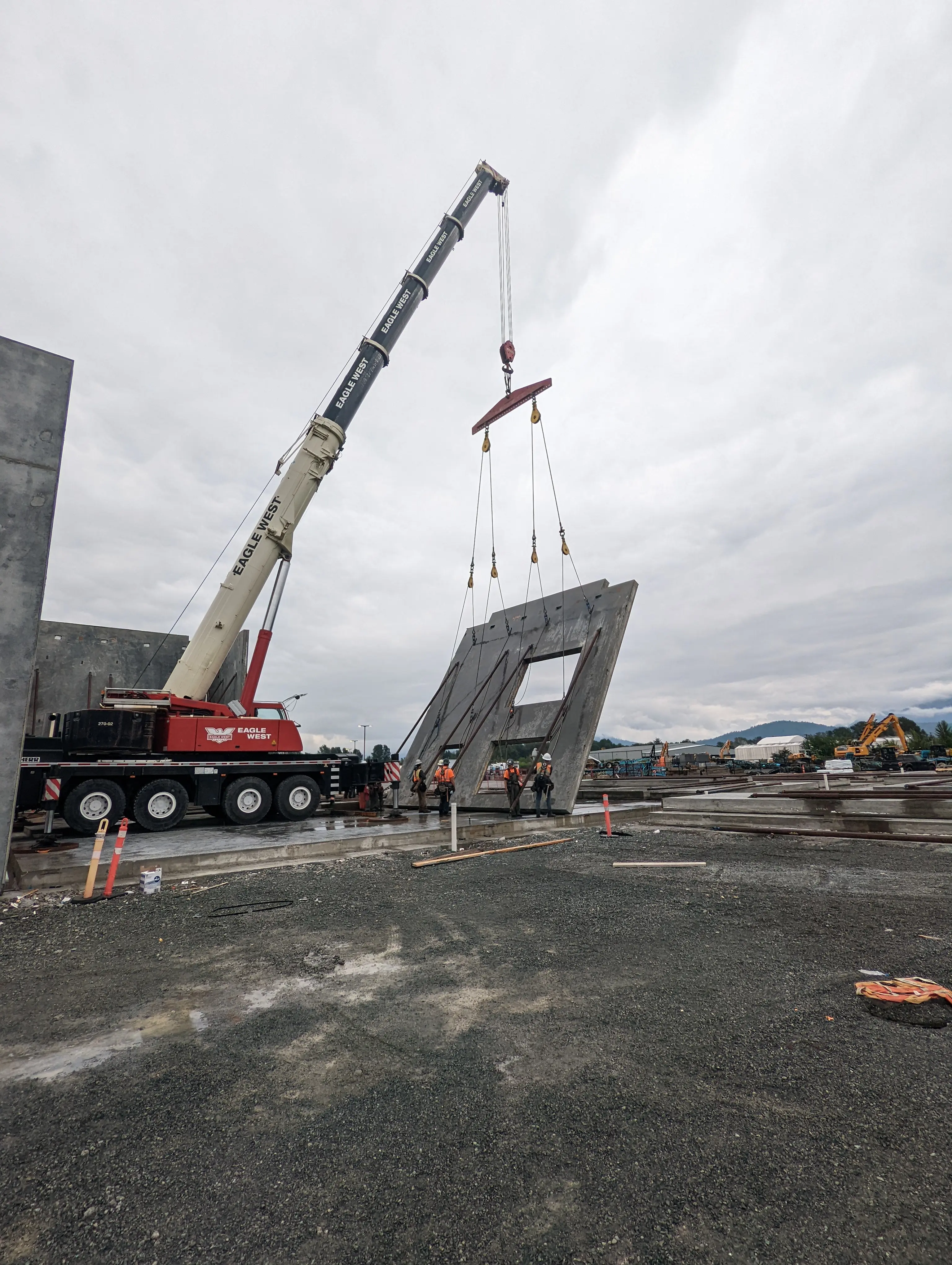 A construction crane lifts a large precast concrete wall panel vertically into place at a building site, with workers guiding the installation.