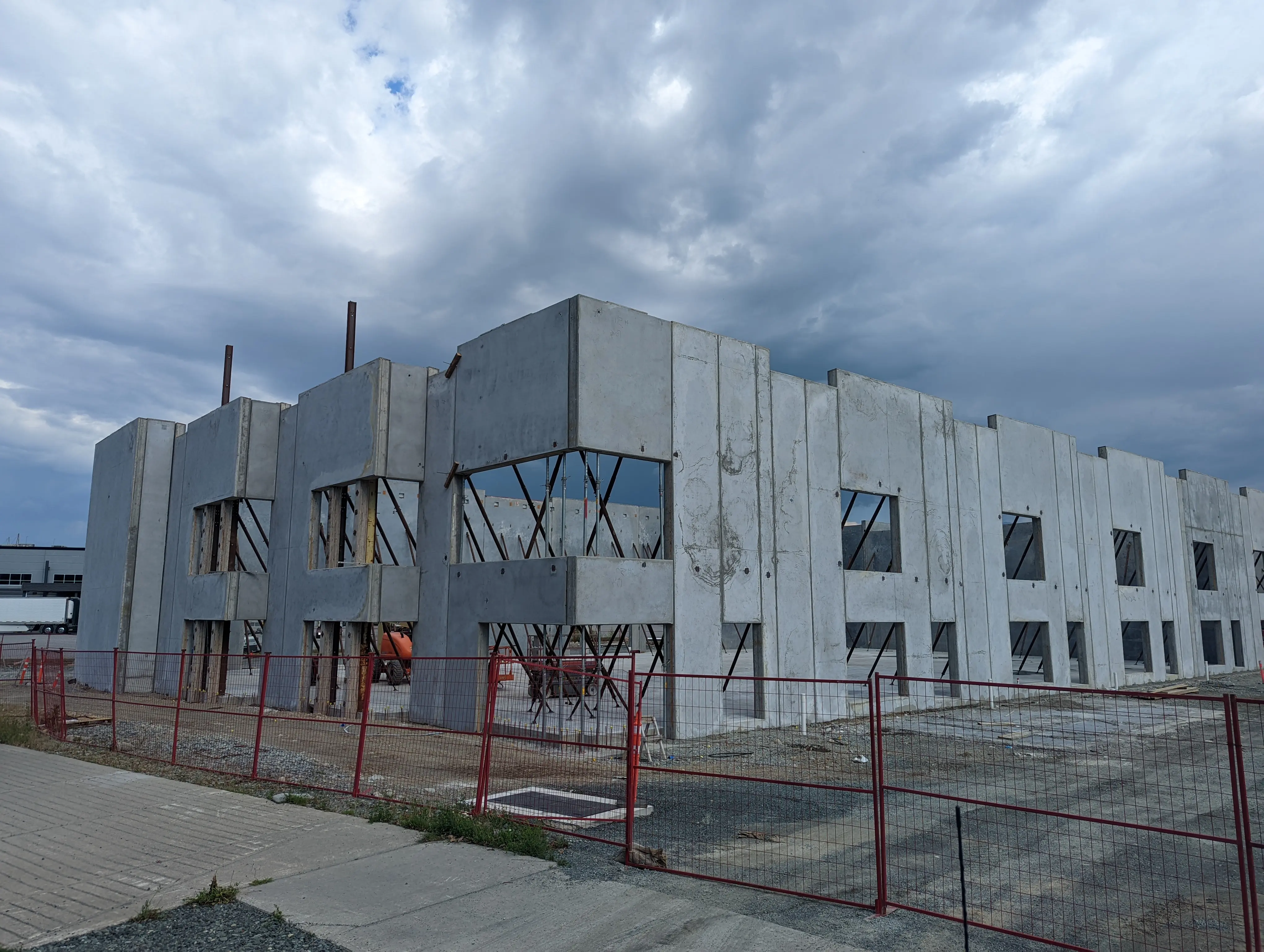 Exterior view of a precast concrete building under construction, with multiple wall panels erected and temporarily braced, surrounded by safety fencing.