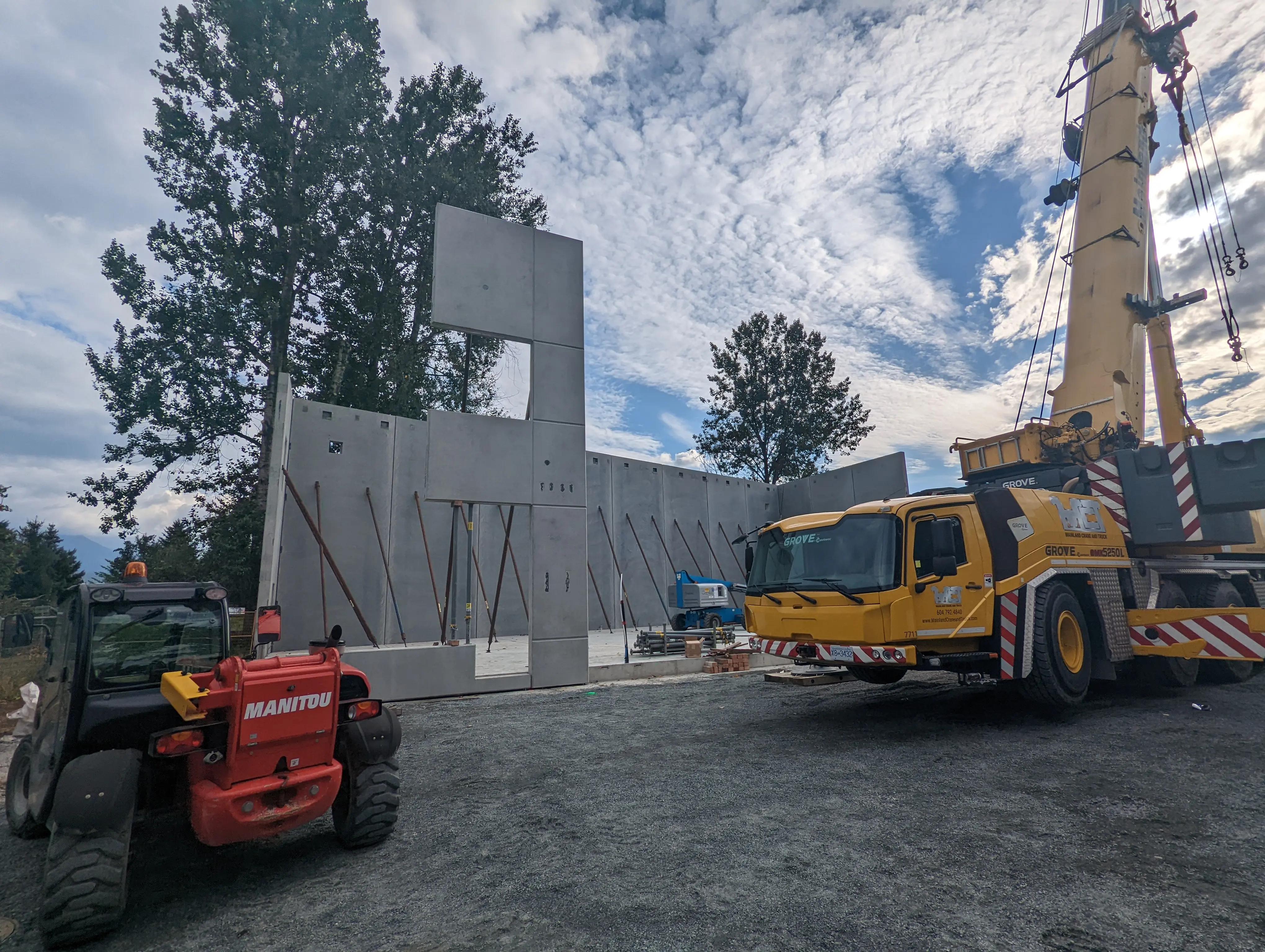 Construction site with a large yellow crane and red forklift positioning precast concrete wall panels into place, supported by temporary steel braces under a cloudy sky.