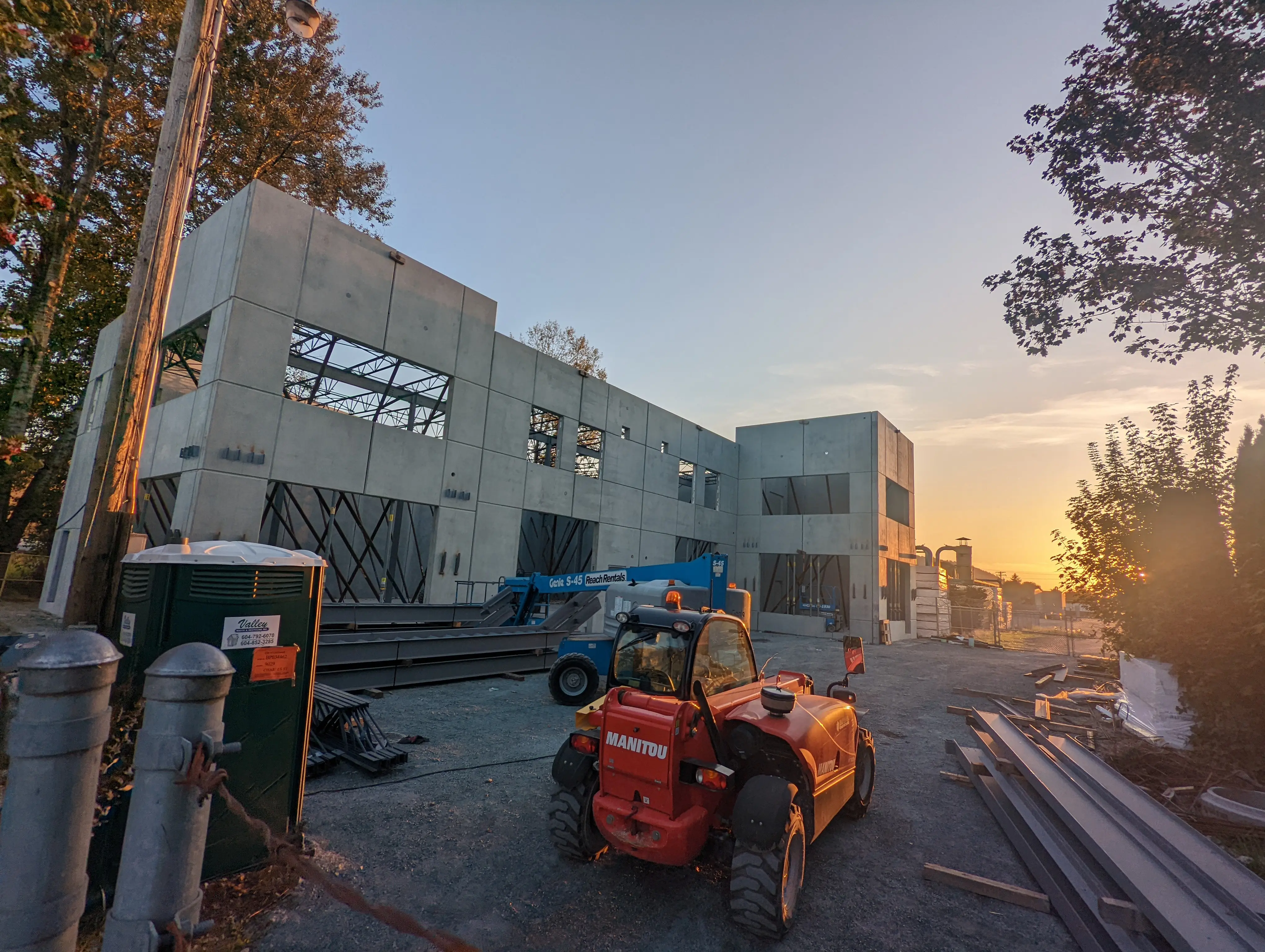 Partially built two-story concrete structure with exposed wall panels and steel bracing, surrounded by construction equipment at sunset.