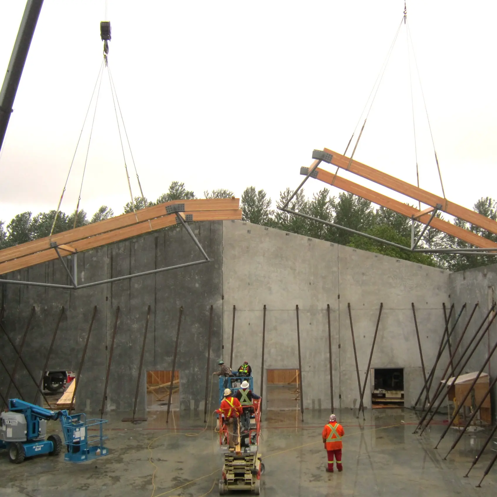 Construction workers and cranes installing large wooden roof trusses atop tall concrete tilt-up walls, with support braces holding the structure in place.