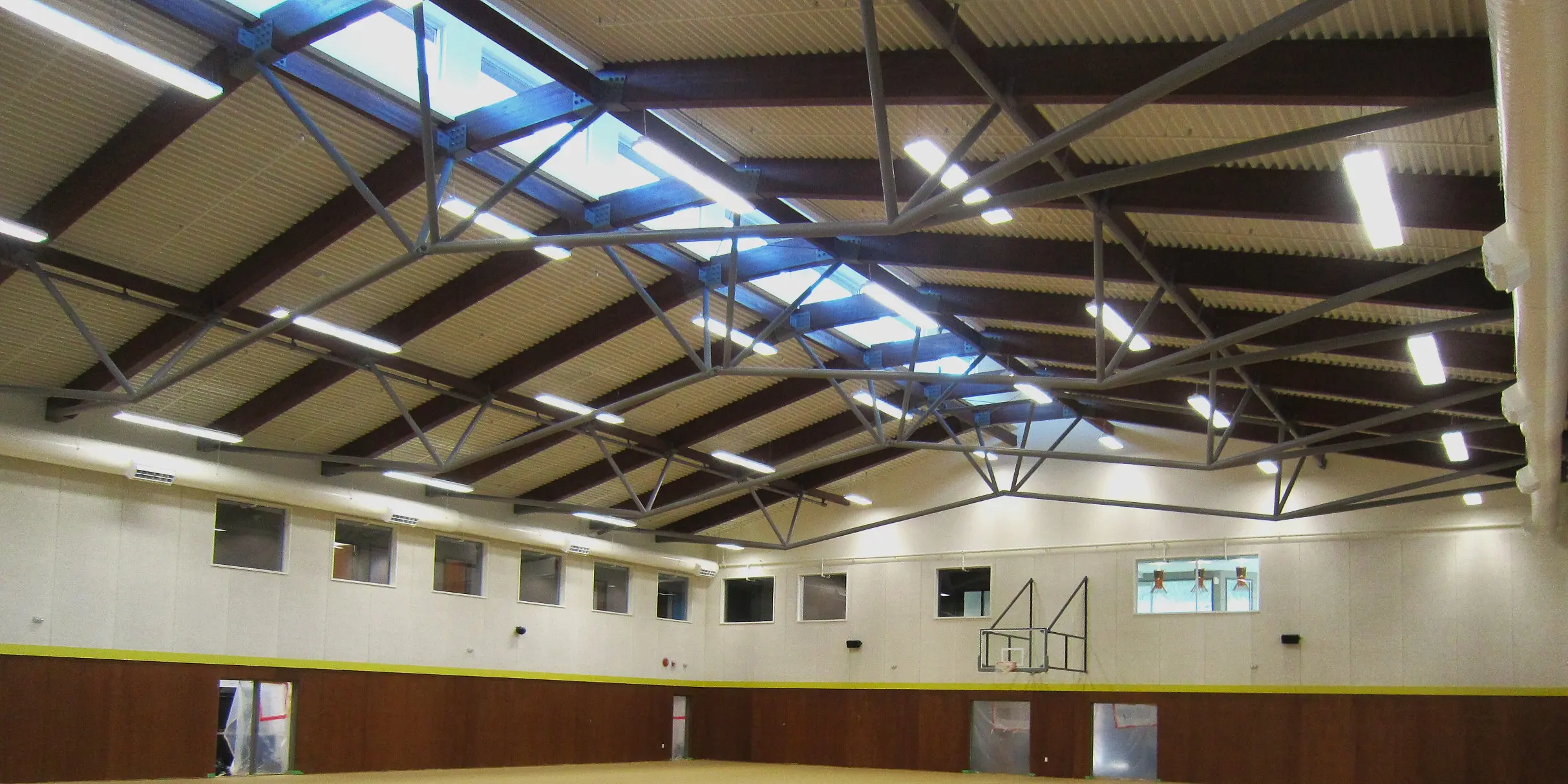 Interior of a gymnasium with exposed steel trusses, skylights, bright overhead lighting, and a basketball hoop mounted on the wall.