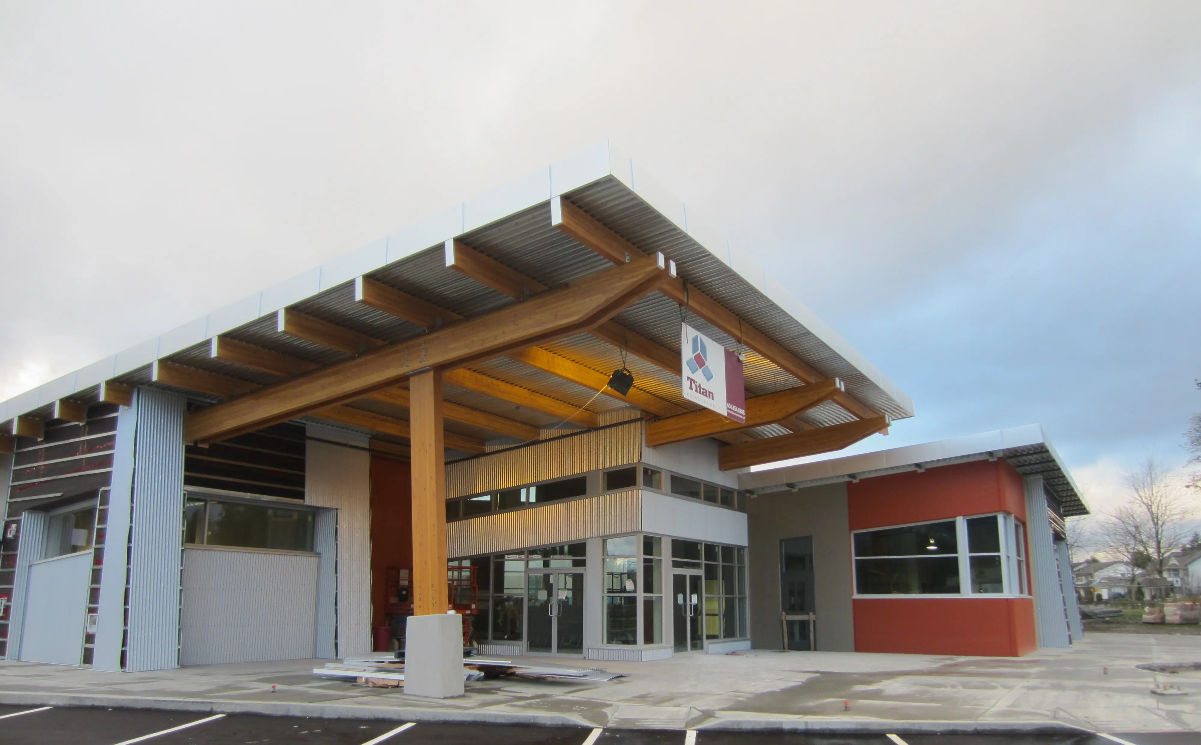 Exterior of a modern commercial building featuring a large timber canopy, metal siding, and glass entrance doors with a Titan Construction sign displayed.