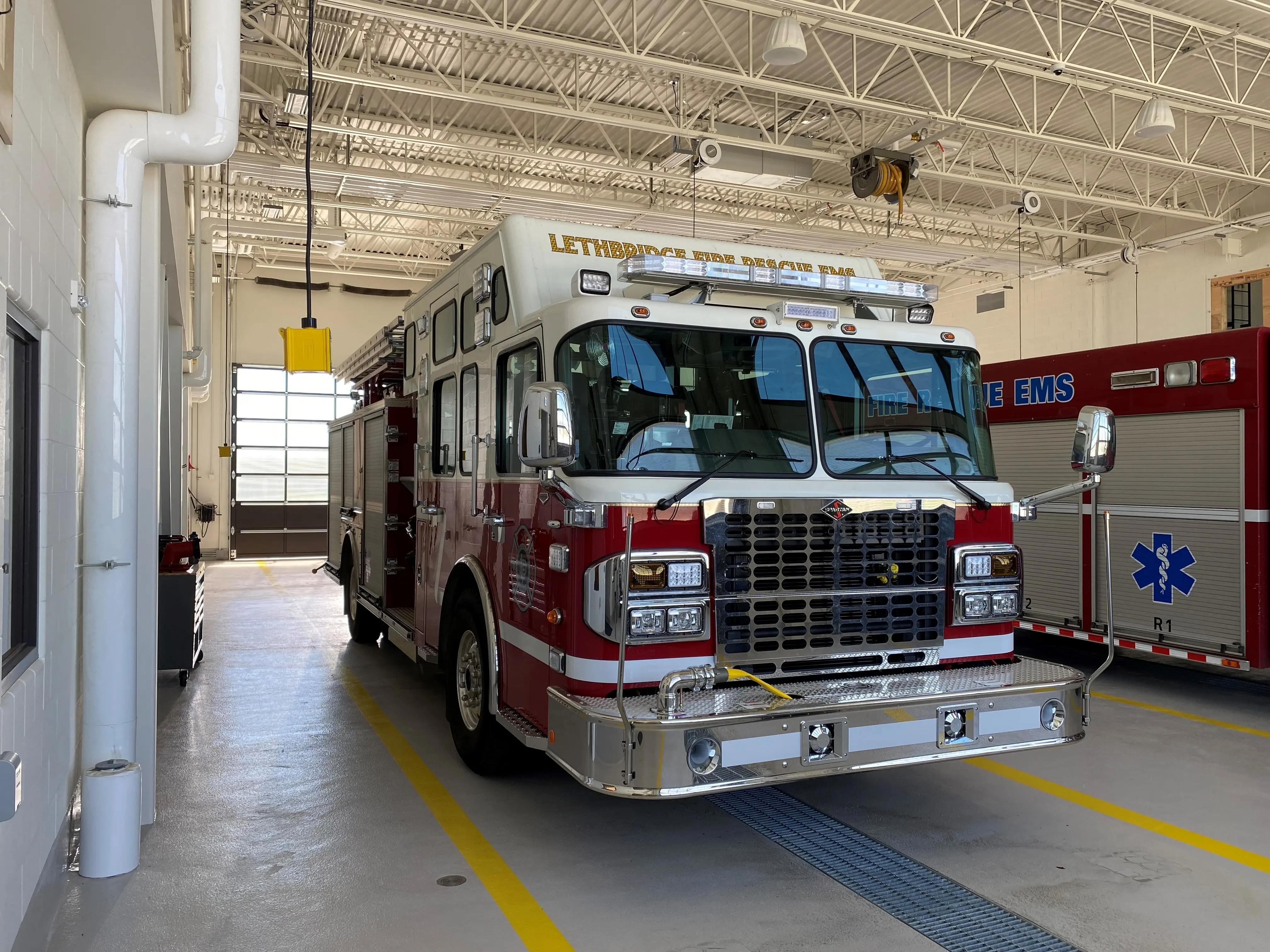 Red fire truck parked inside a bright, modern fire hall garage with white walls and overhead piping.