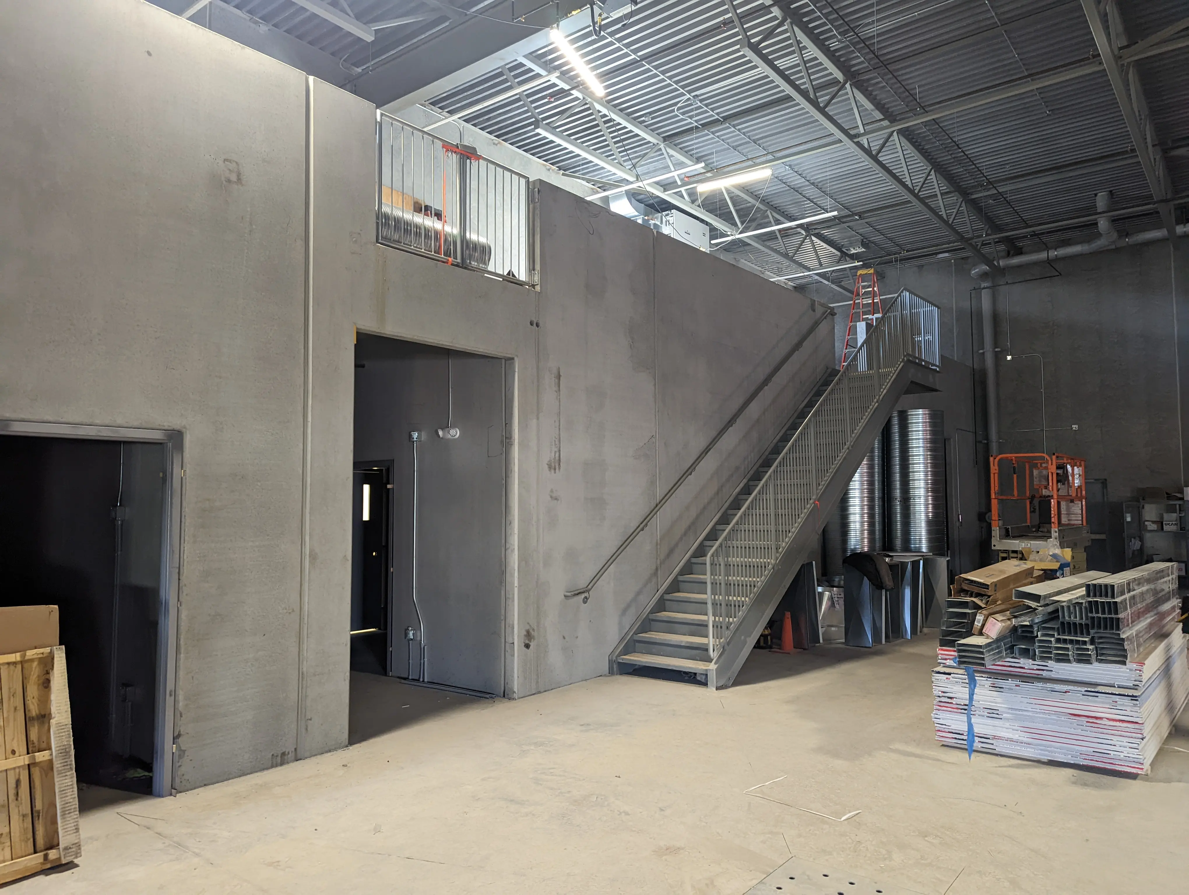 A partially completed interior of a fire station, showing a concrete structure with a staircase leading to a mezzanine. Metal framing, ductwork, and construction materials are organized on the floor.