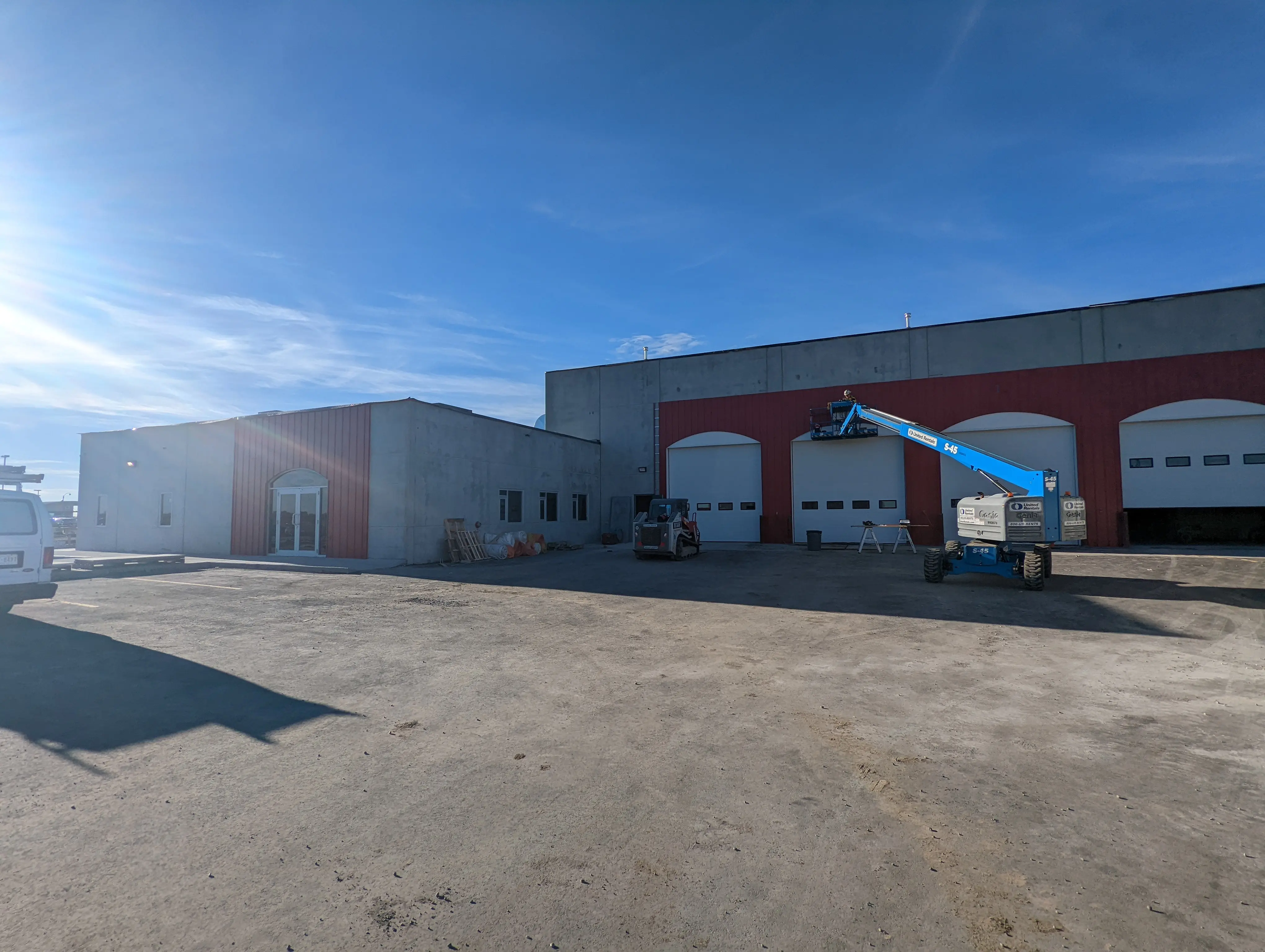 A wide-angle view of the fire station exterior with large bay doors, red siding accents, and construction equipment including a blue boom lift positioned near the building.