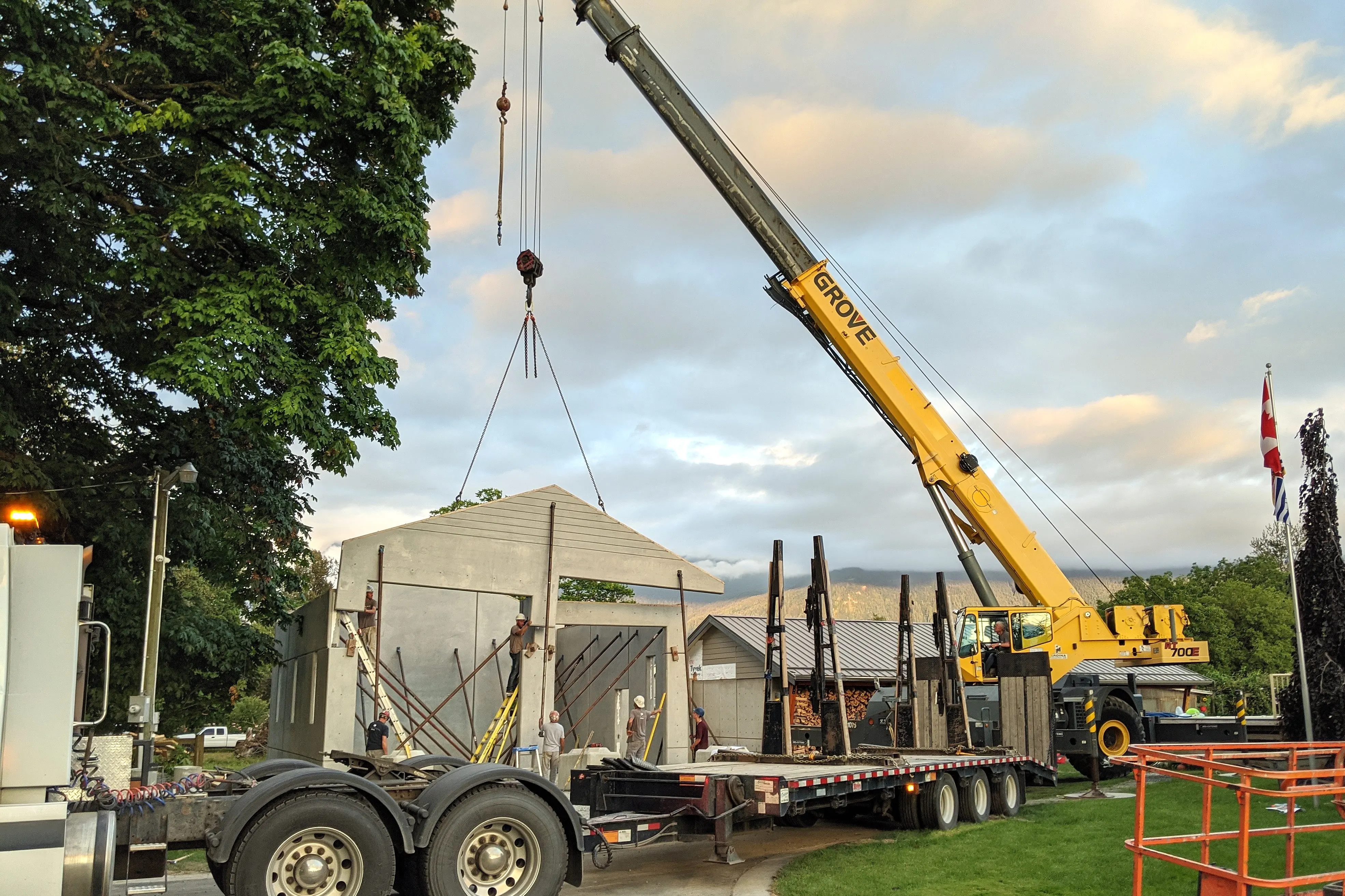 A yellow Grove crane lifting and installing a large precast concrete wall panel while workers guide it into place, with trees and a Canadian flag visible in the background.