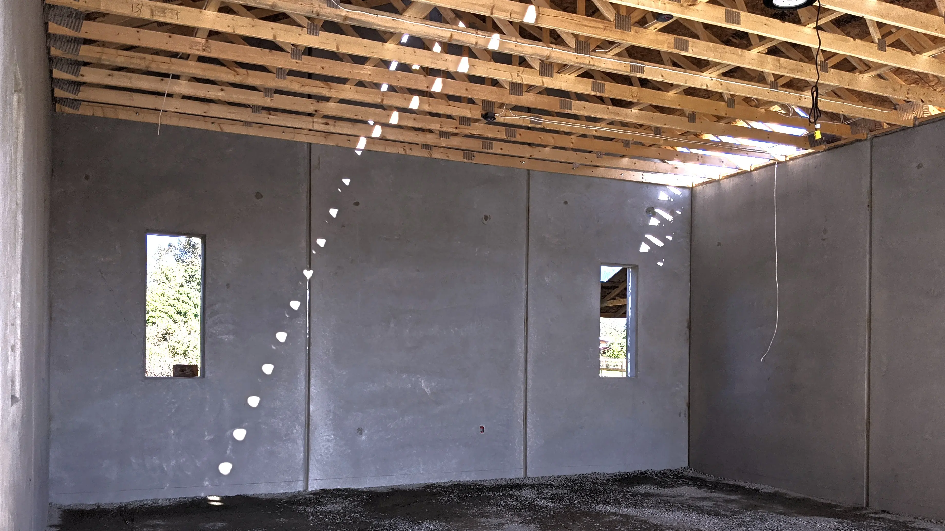 Interior view of a building under construction, showing smooth precast concrete walls with two vertical windows and exposed wooden roof trusses above.