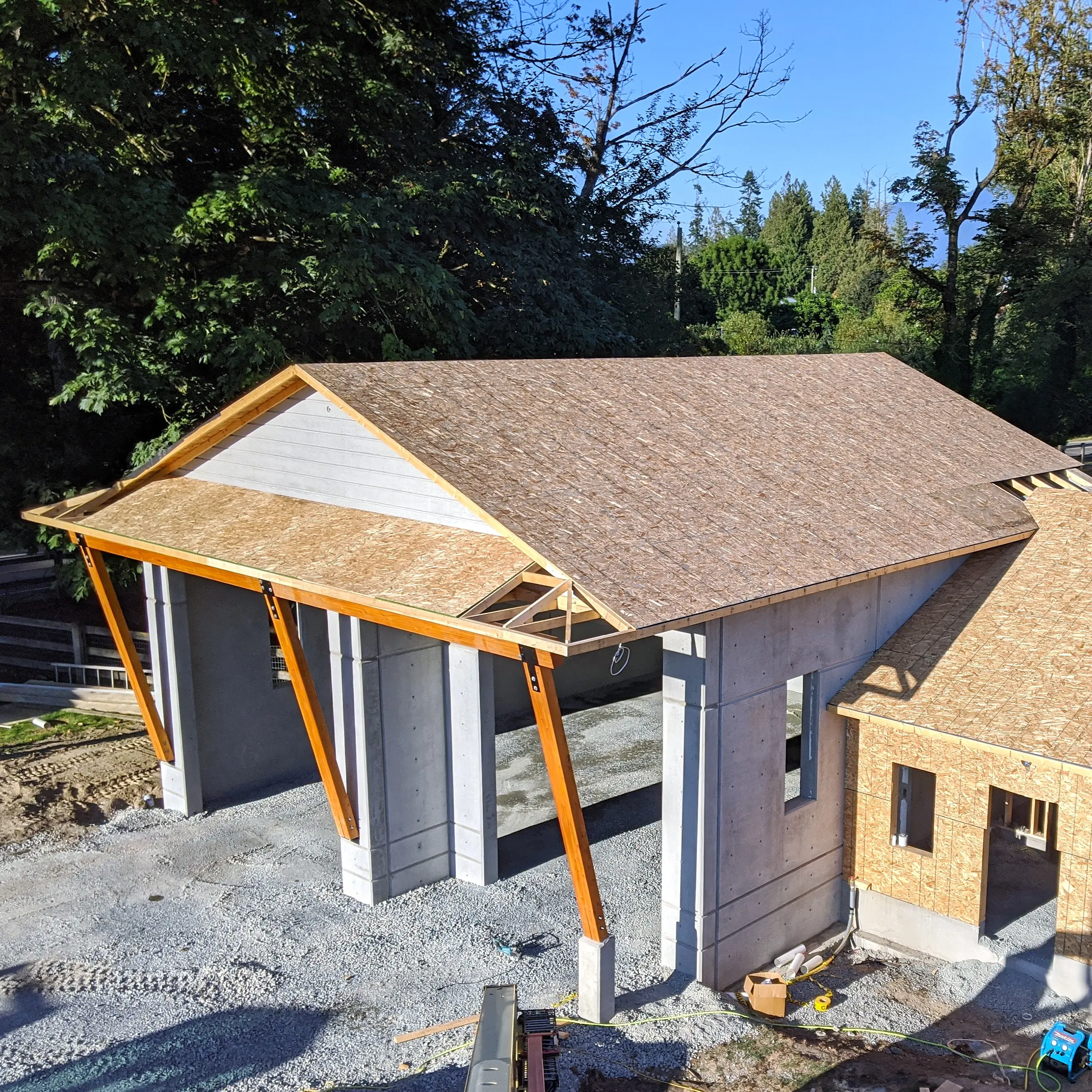 A partially constructed building with precast concrete walls and slanted wooden roof framing, supported by angled timber columns, surrounded by gravel and trees.