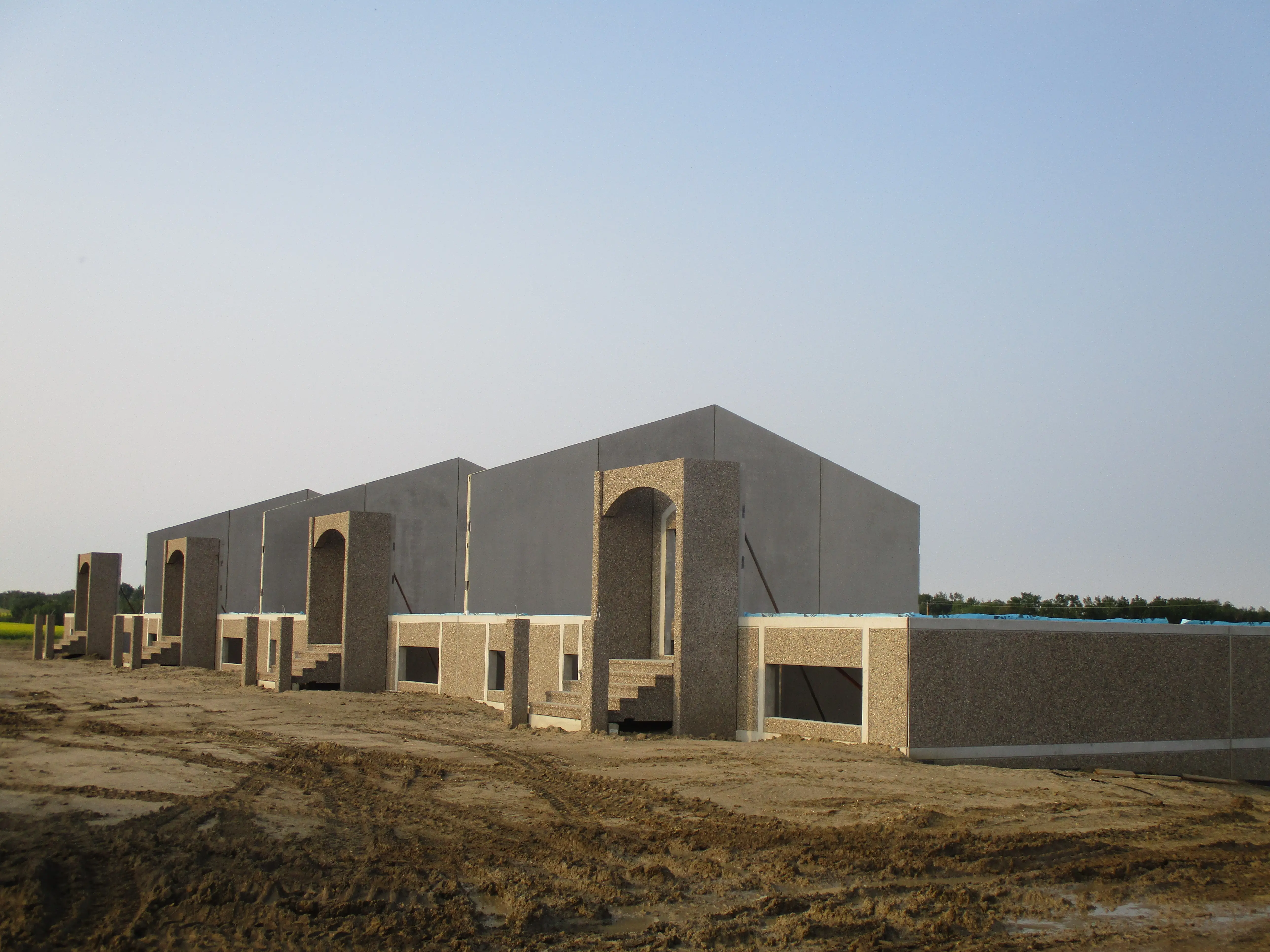 Exterior construction progress of a large building with precast concrete walls and exposed aggregate finish, standing on a dirt site.