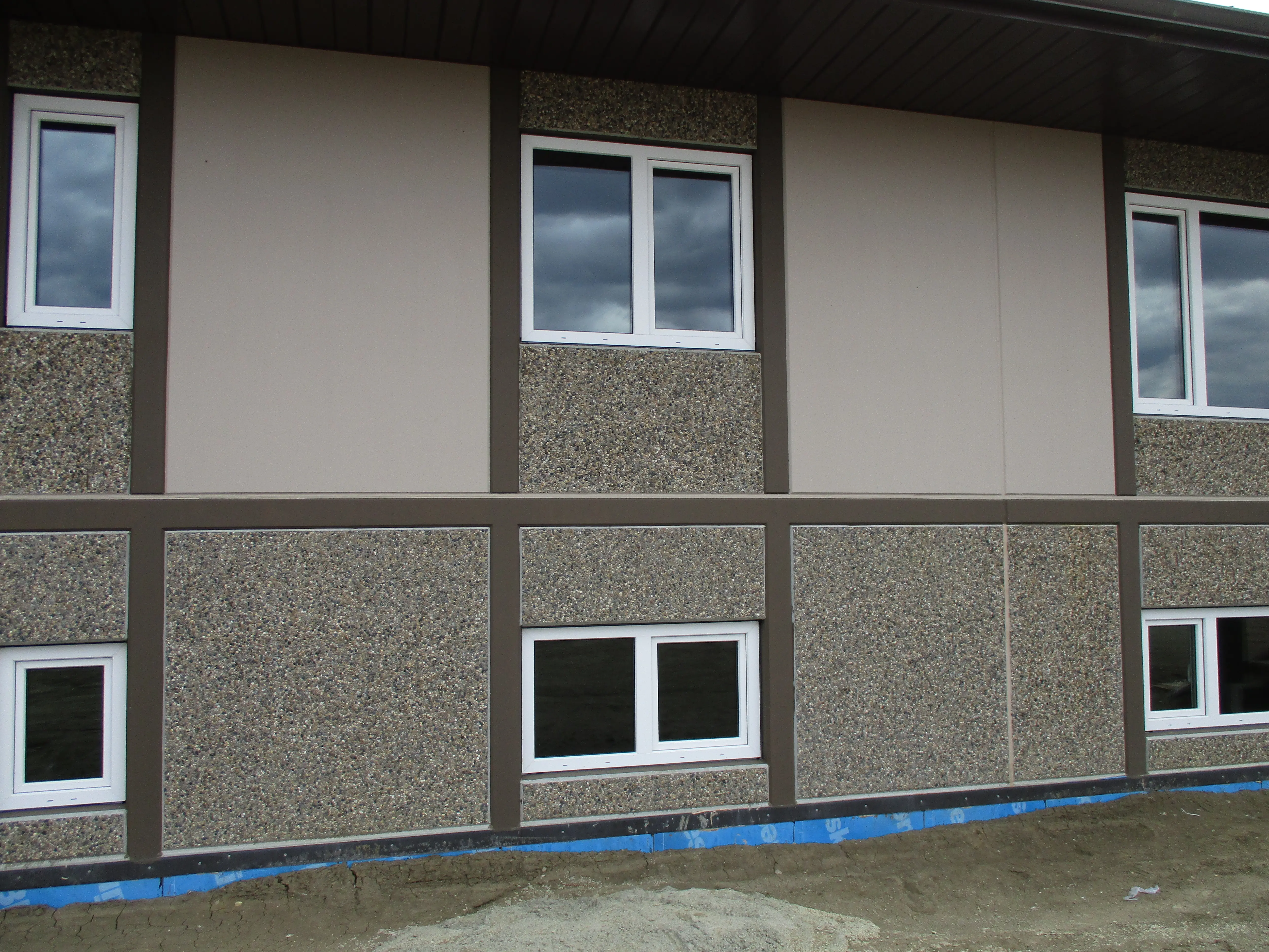 Exterior wall of a completed building featuring pebble-textured concrete panels, brown framing, and multiple white-framed windows.