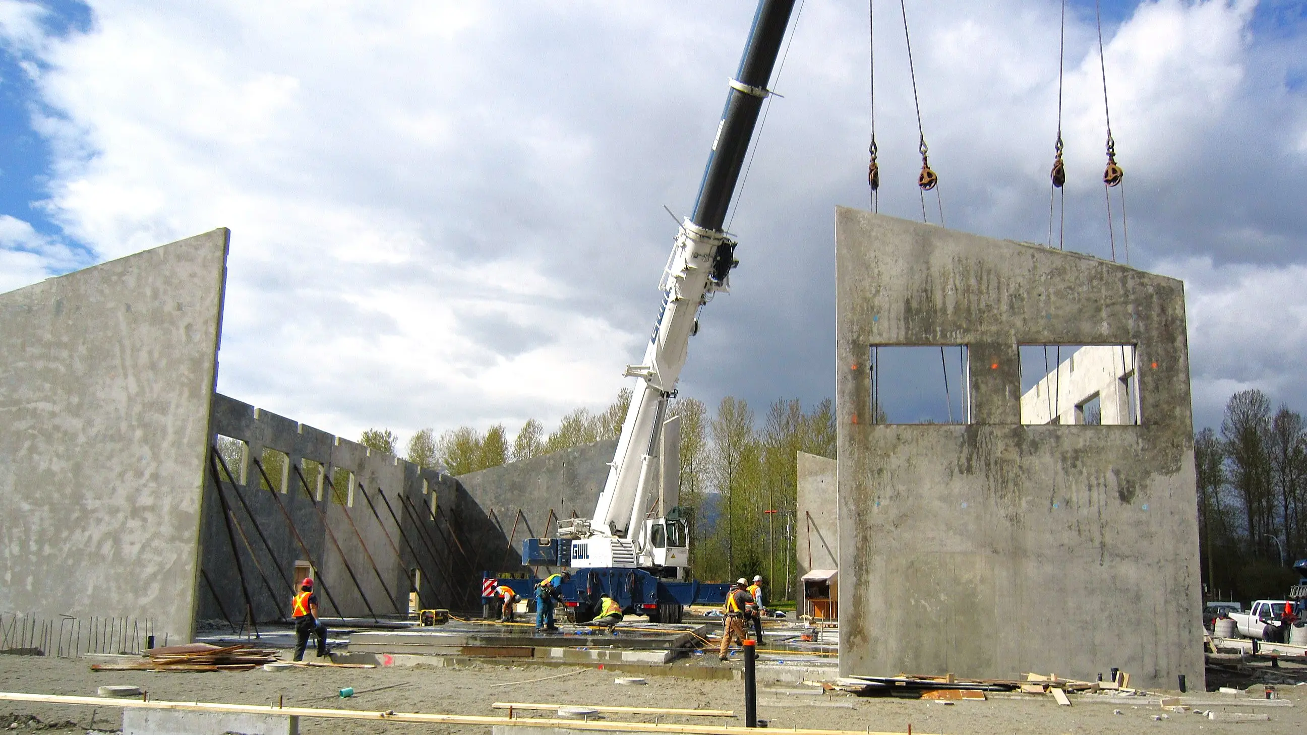 Construction workers and cranes installing large wooden roof trusses atop tall concrete tilt-up walls, with support braces holding the structure in place.