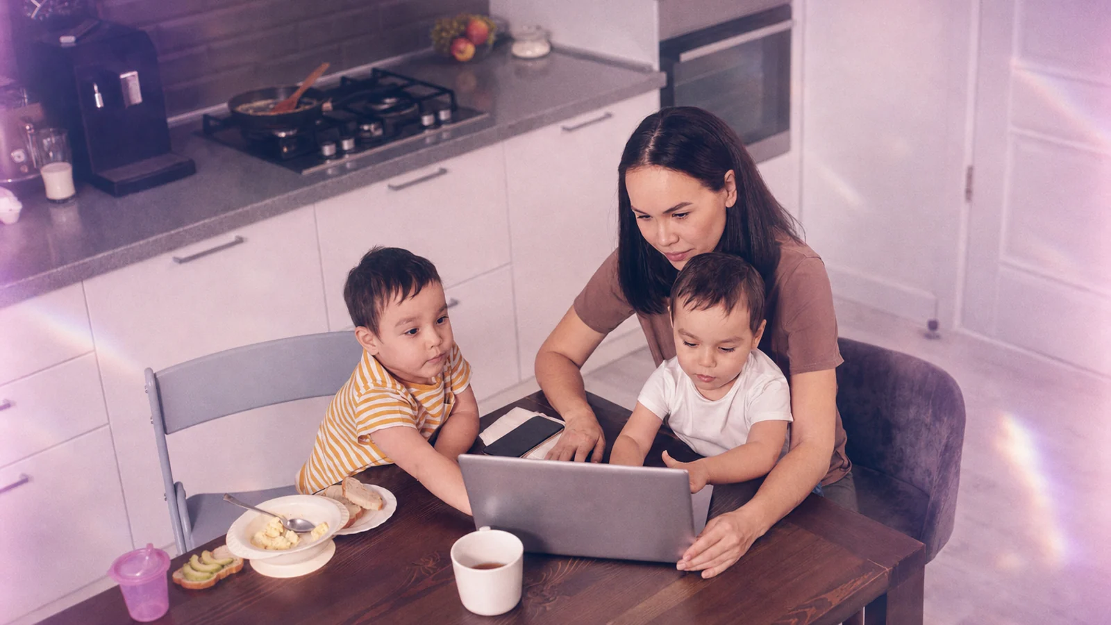 A parent using a laptop at the kitchen table with two young children
