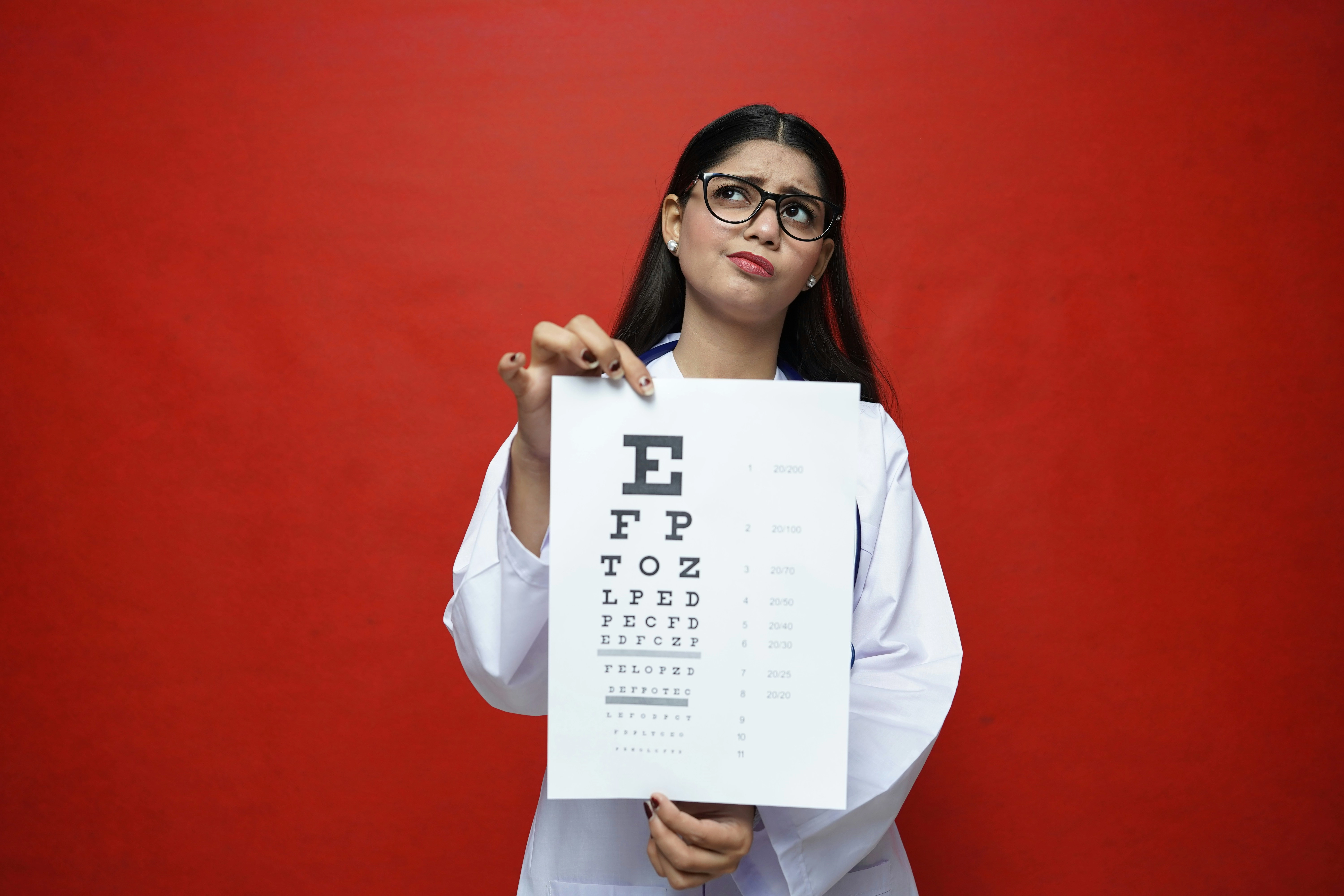 Eye care professional holding a Snellen eye chart against a red background while wearing glasses and a white coat