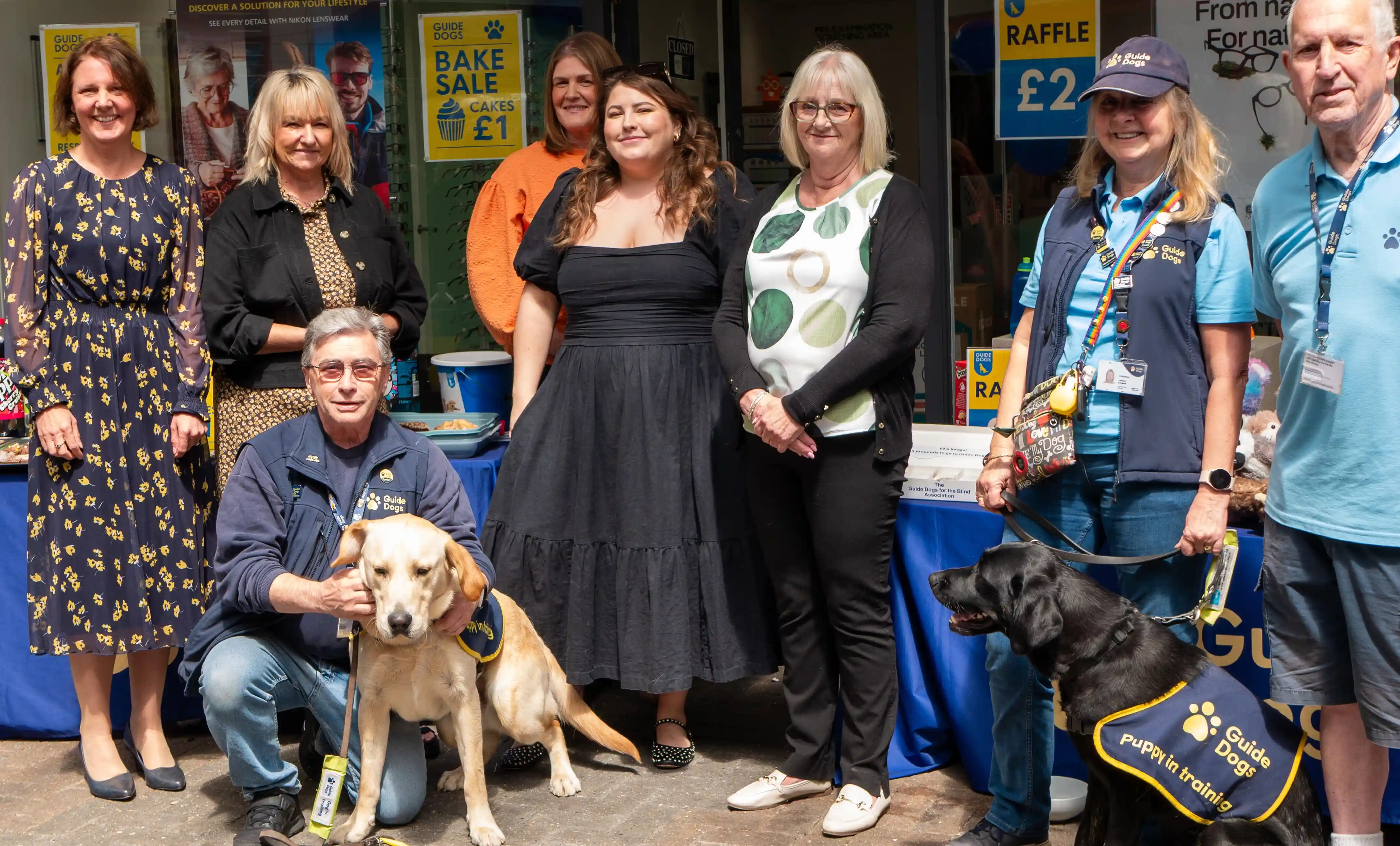 Group of community members and Guide Dogs volunteers posing outside an optician practice with guide dogs and fundraising displays during a charity event