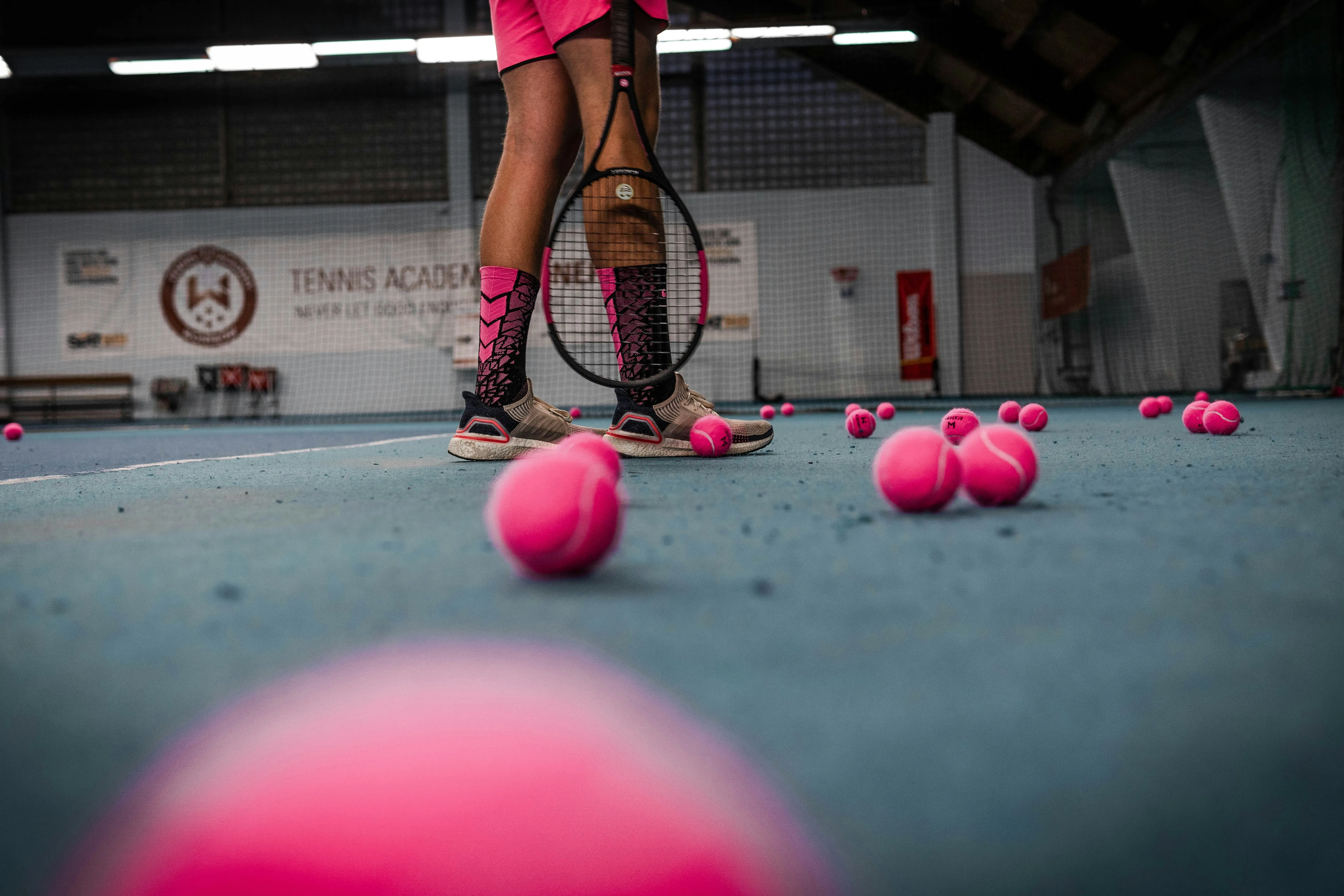 Tennis player standing on an indoor court with a racket surrounded by bright pink tennis balls during a training session