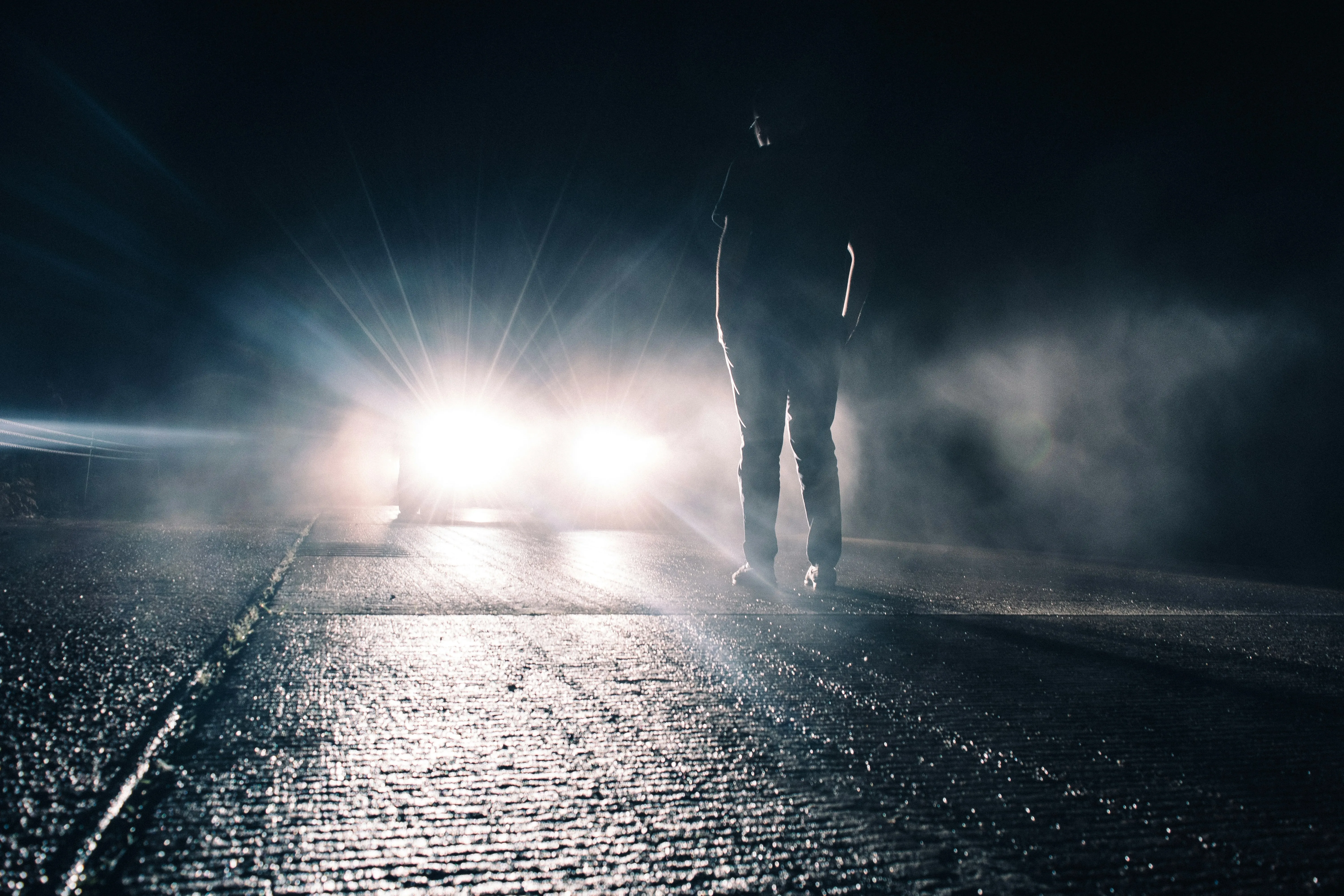 Person standing on a dark road at night facing bright vehicle headlights, illustrating the challenges of glare and reduced visibility after dark