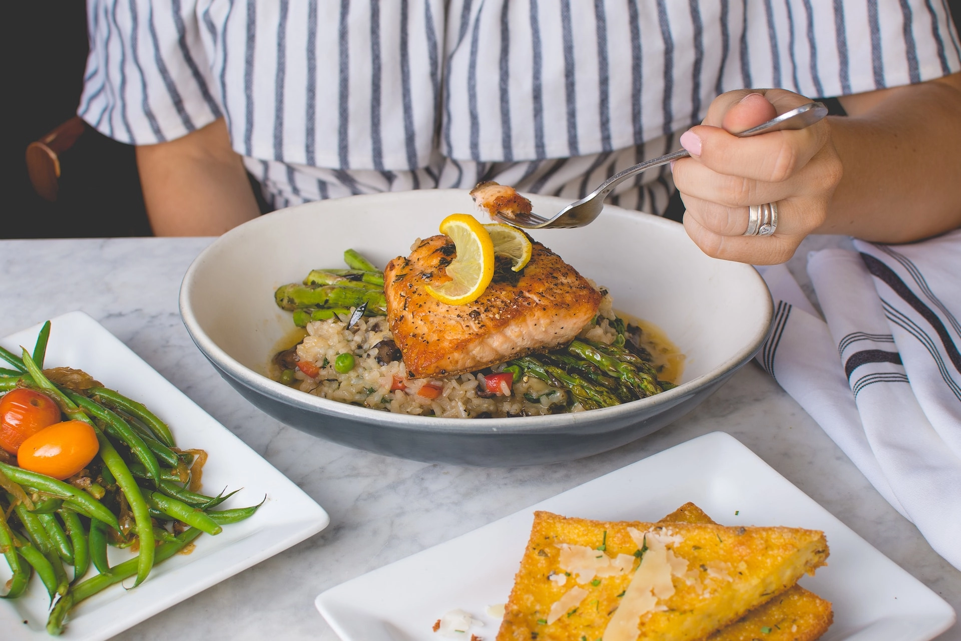 Person enjoying a healthy meal featuring grilled salmon, asparagus, rice, and fresh vegetables at a restaurant table