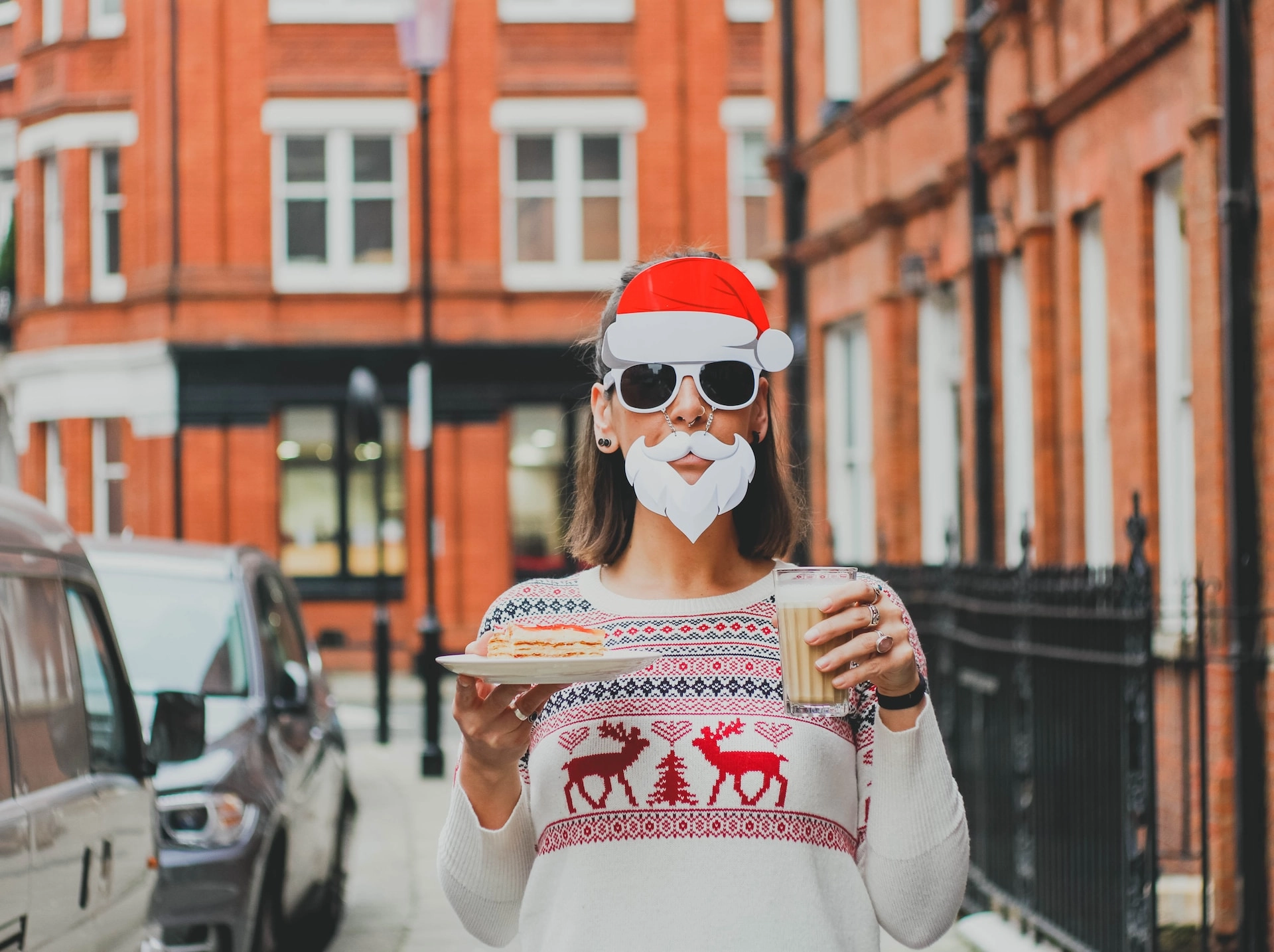 Person wearing festive Christmas-themed glasses and a holiday jumper while enjoying a drink and dessert on a city street