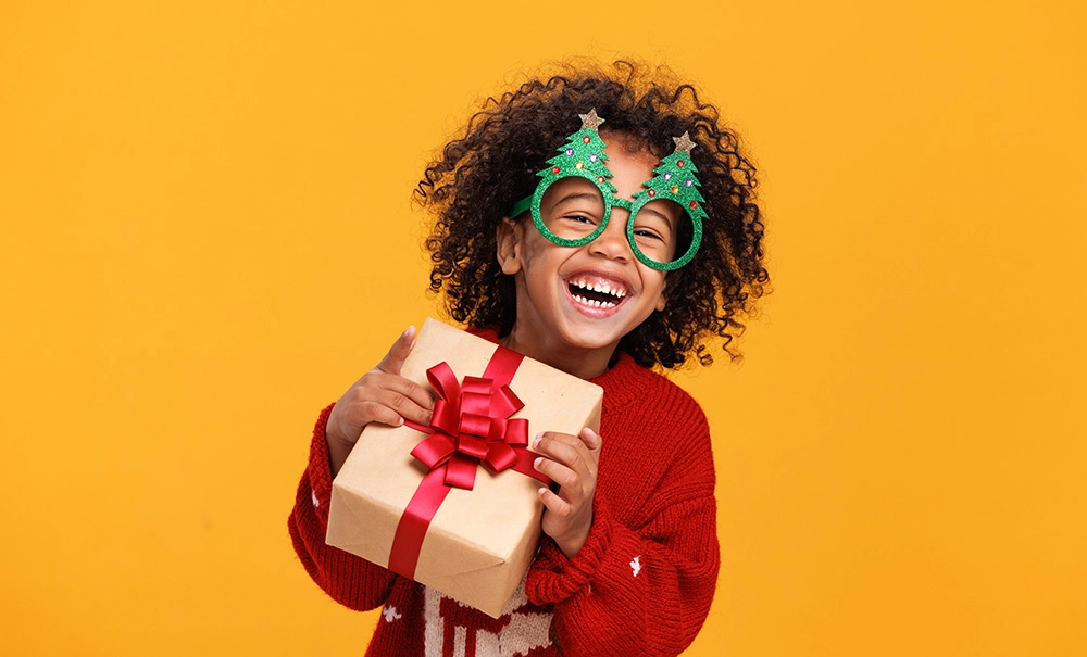Smiling child wearing Christmas tree novelty glasses and holding a wrapped gift.