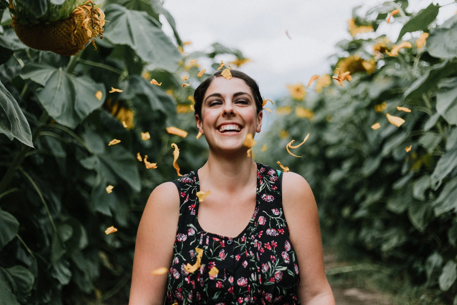 Smiling woman standing among tall sunflowers as petals fall around her, celebrating outdoor wellbeing and enjoying nature