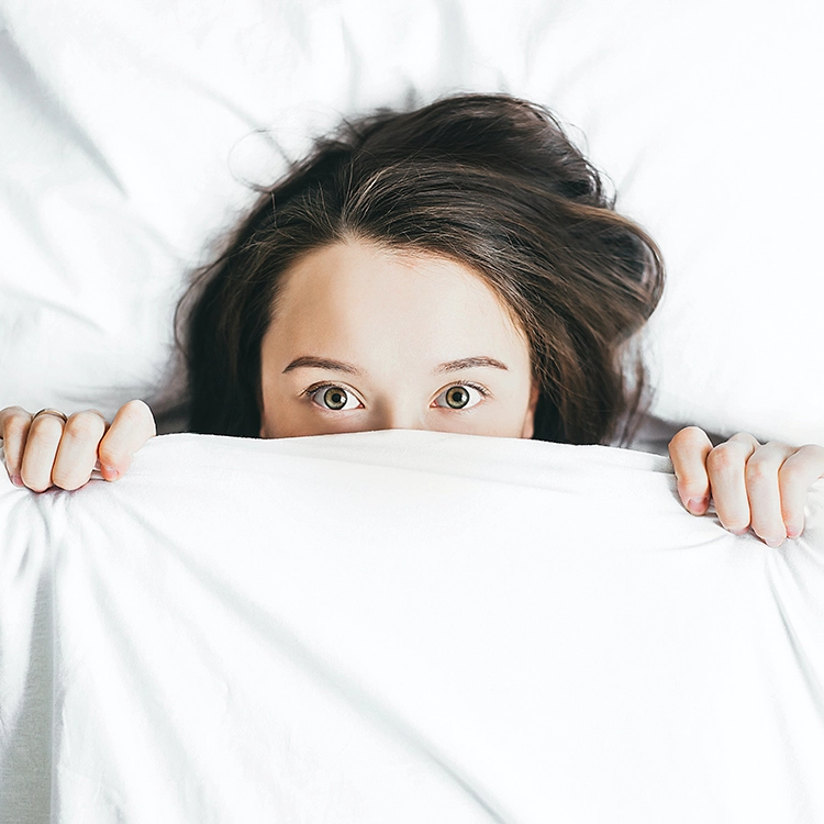 Woman peeking over a white blanket with wide-open eyes, highlighting eye awareness, vision health, and the importance of regular eye care