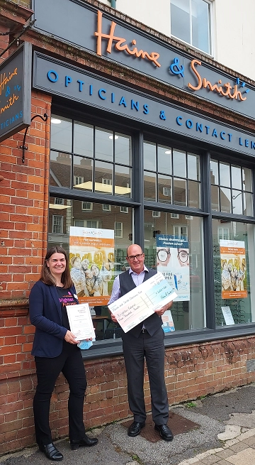 Two people standing outside Haine & Smith Opticians holding a donation cheque and certificate in front of the practice storefront