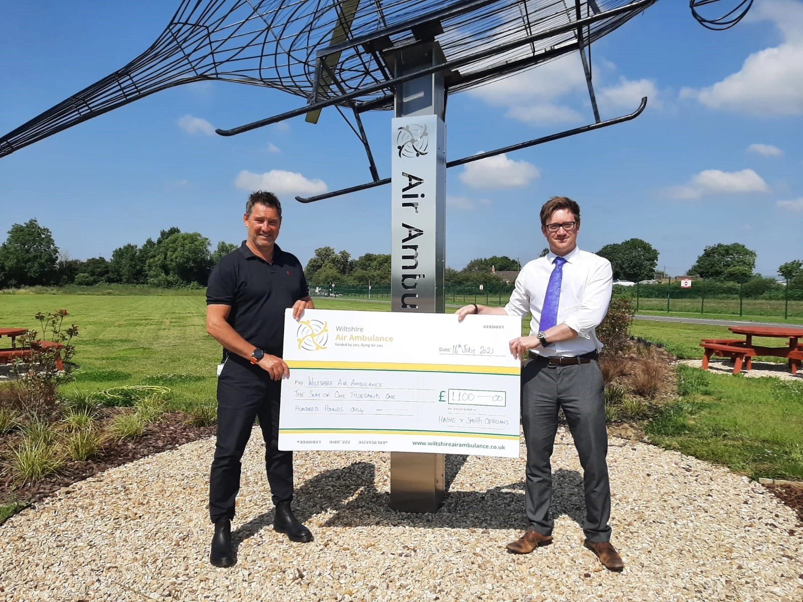 Two individuals presenting a charitable donation cheque to Wiltshire Air Ambulance beside an air ambulance-themed sculpture in an outdoor setting