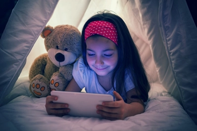 Young girl using a tablet inside a blanket fort at night, illuminated by the screen's glow, with a teddy bear beside her