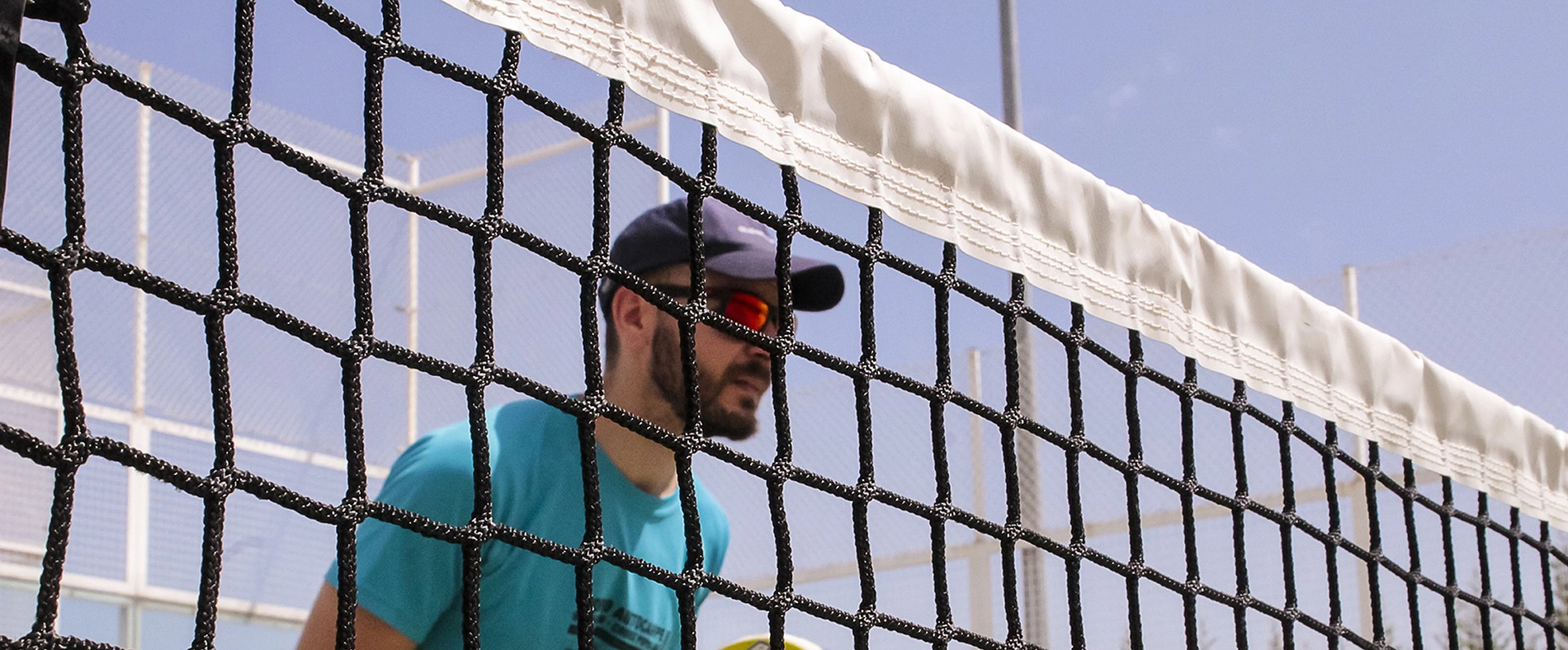 Person wearing sports sunglasses standing behind a court net during an outdoor racket sport match on a sunny day