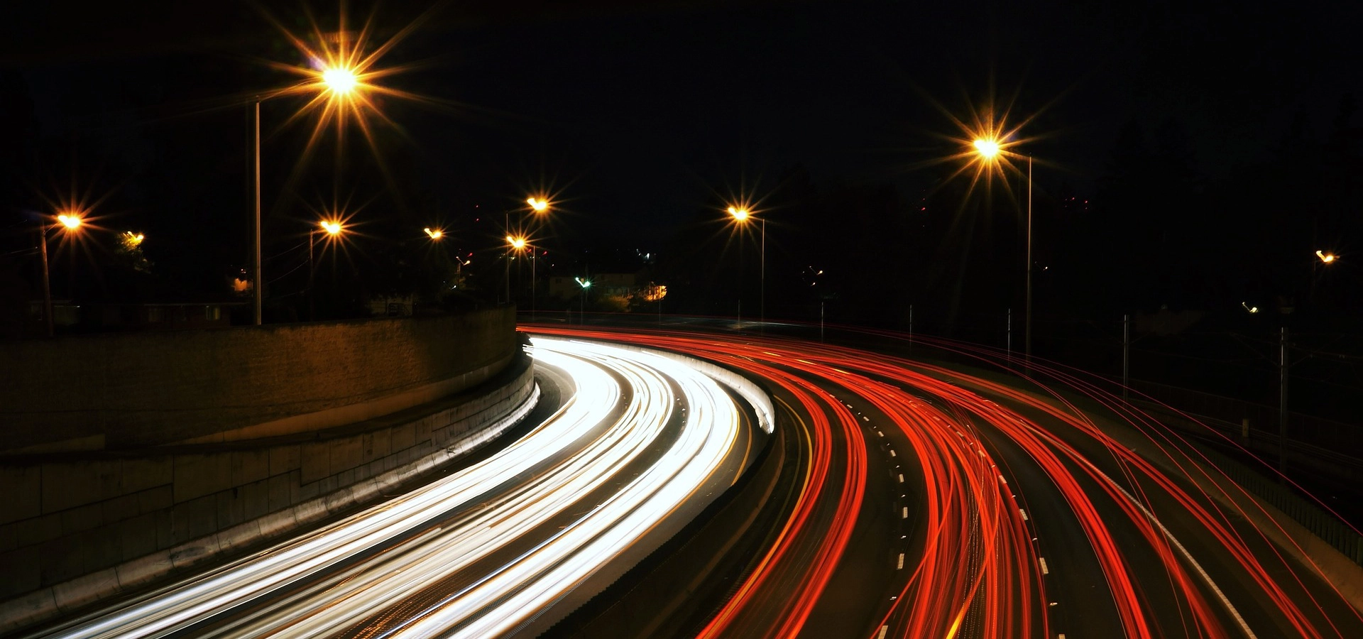 Night-time motorway with bright vehicle light trails and streetlights creating starburst effects along a curved roadway