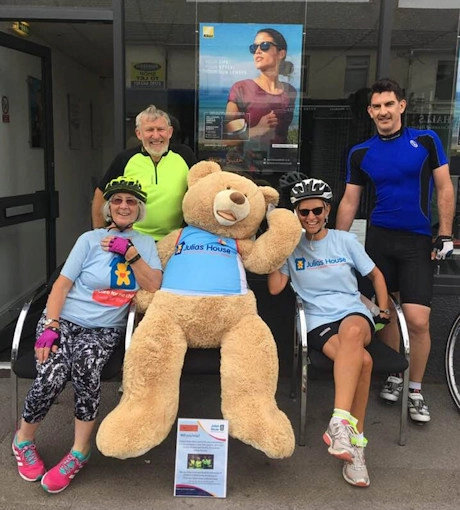 Group of charity cyclists posing with a large teddy bear outside an opticians practice during a fundraising cycling event