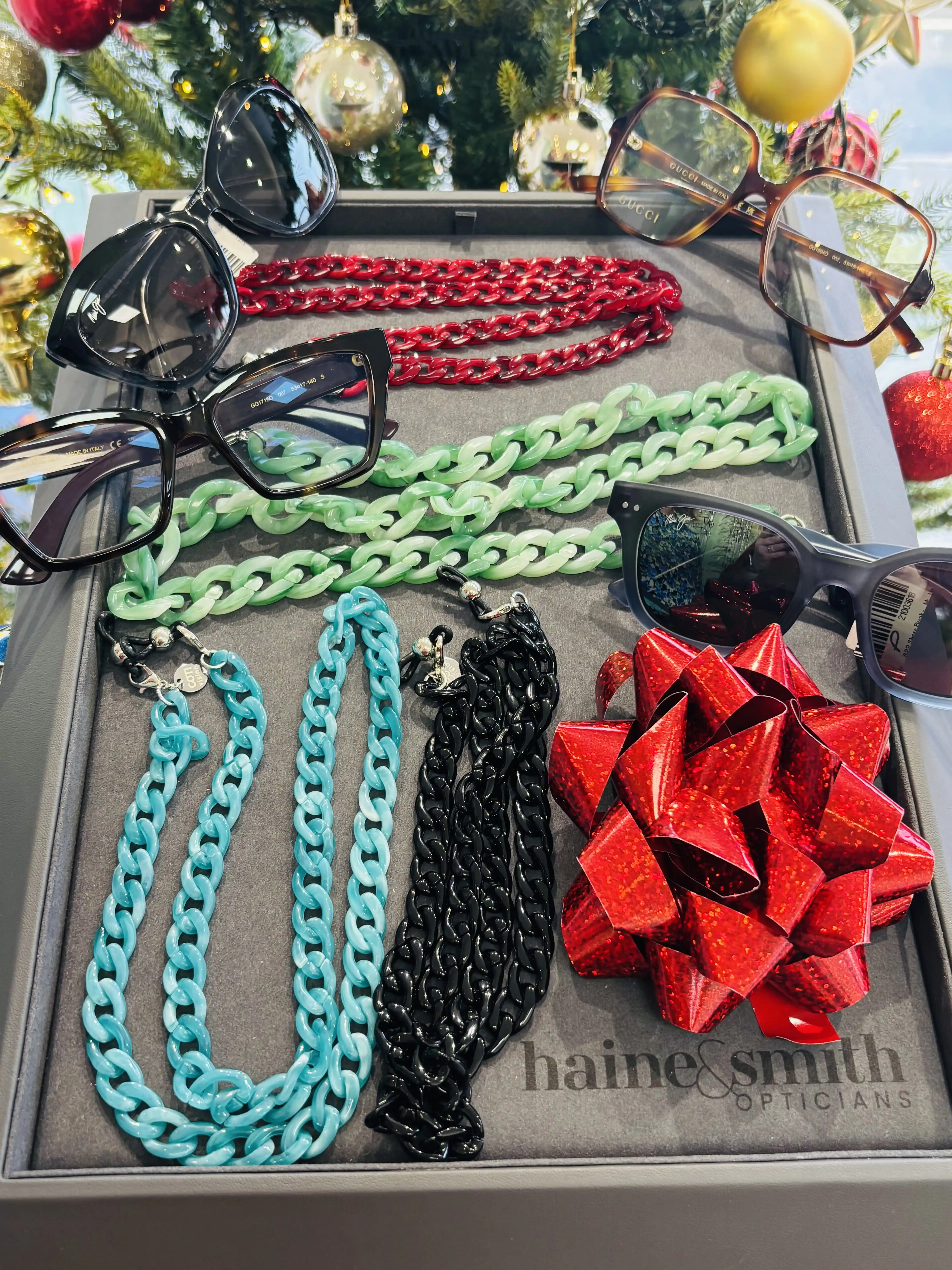 Display of colourful eyewear chains, designer glasses, and sunglasses arranged in a gift tray beneath a decorated Christmas tree