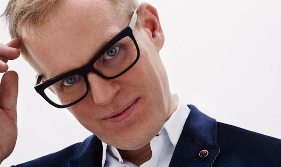 Man wearing bold black rectangular eyeglass frames and a navy blazer in a close-up portrait