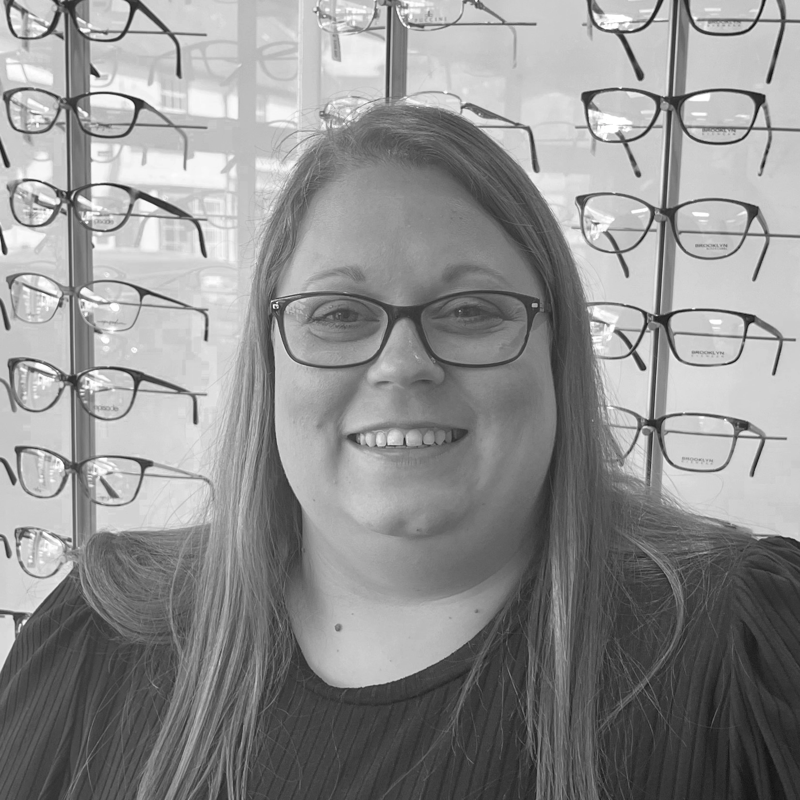 Smiling optician wearing round-frame glasses standing in front of a display wall showcasing a wide selection of prescription eyewear frames