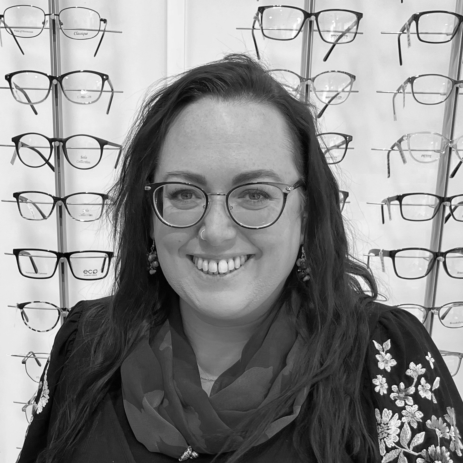 Smiling optician wearing round-frame glasses standing in front of a display wall showcasing a wide selection of prescription eyewear frames