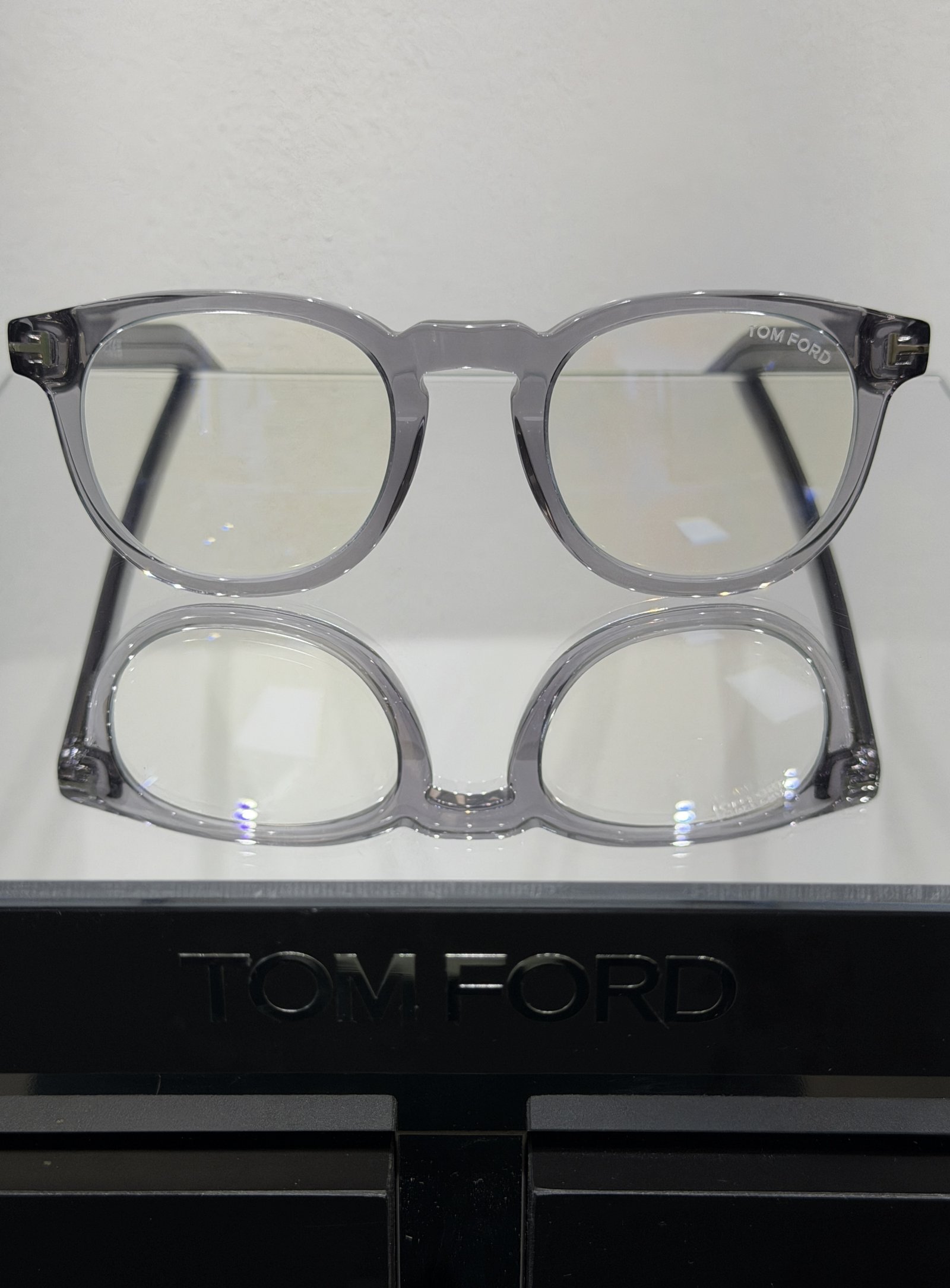 Tom Ford transparent grey eyeglass frames displayed on a branded optical stand