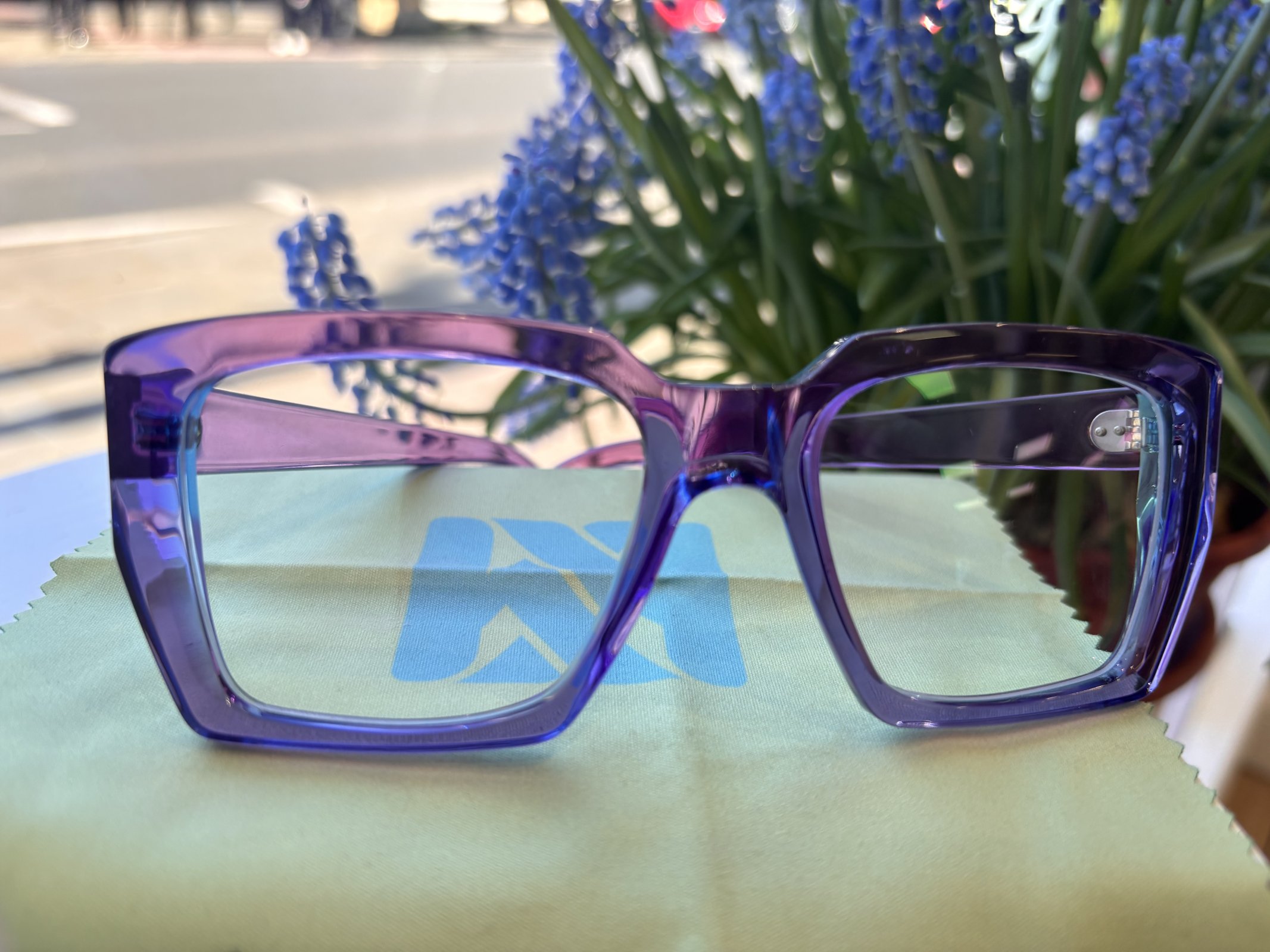 Bold translucent purple eyeglass frames displayed outdoors with blue flowers in the background
